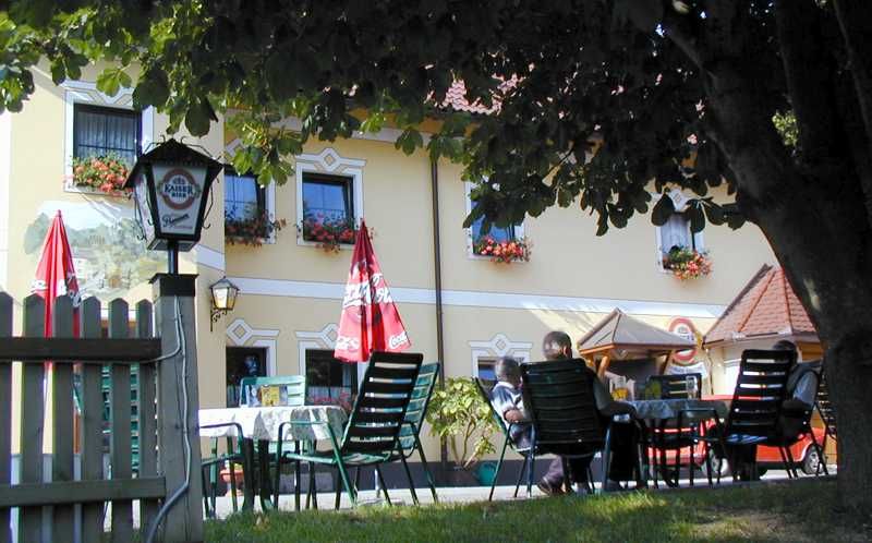 A beer garden with tables and chairs under a tree in front of a yellow building with flower boxes.