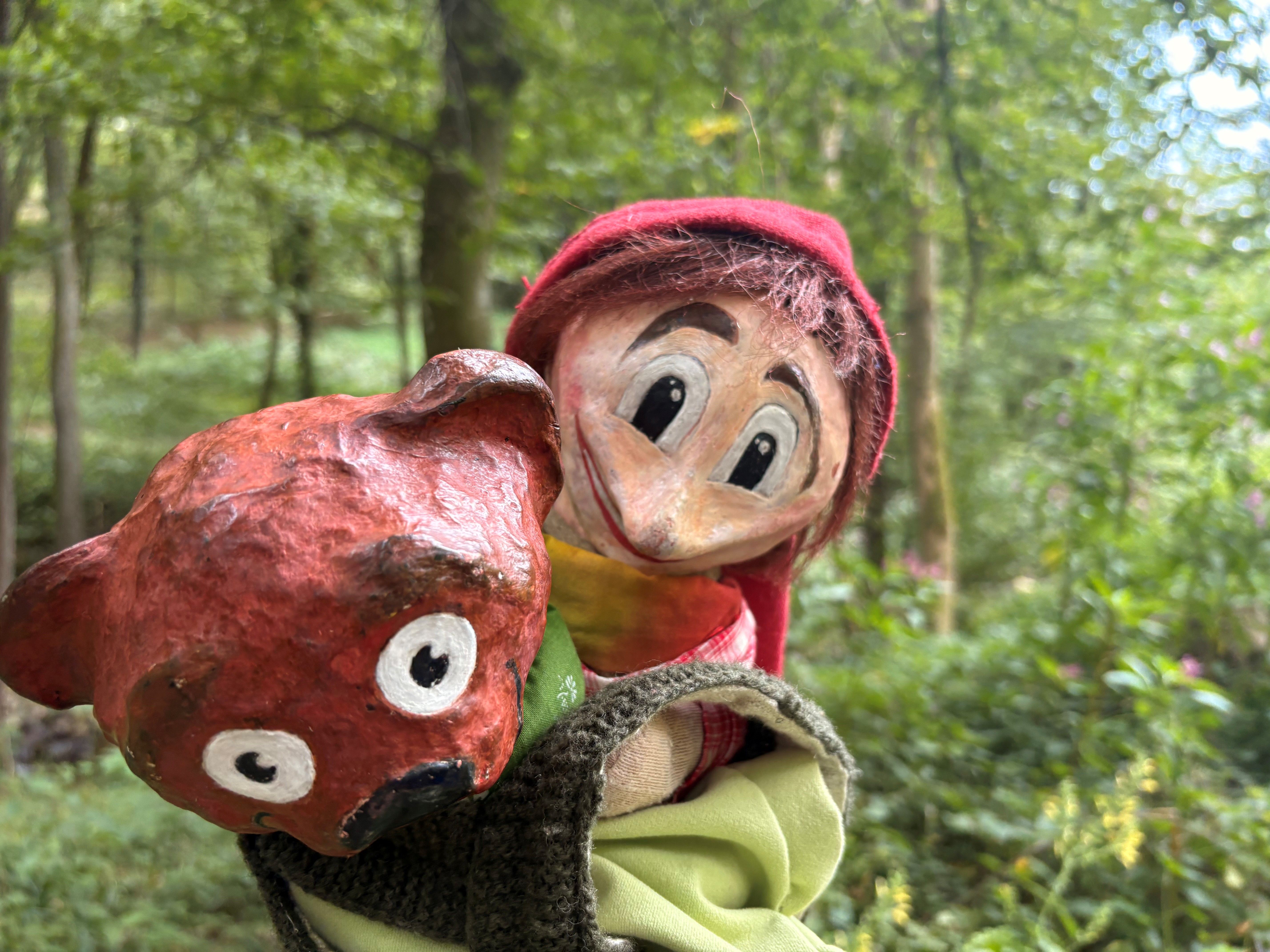 A hand puppet wearing a red hat and holding a red bear in a forest.