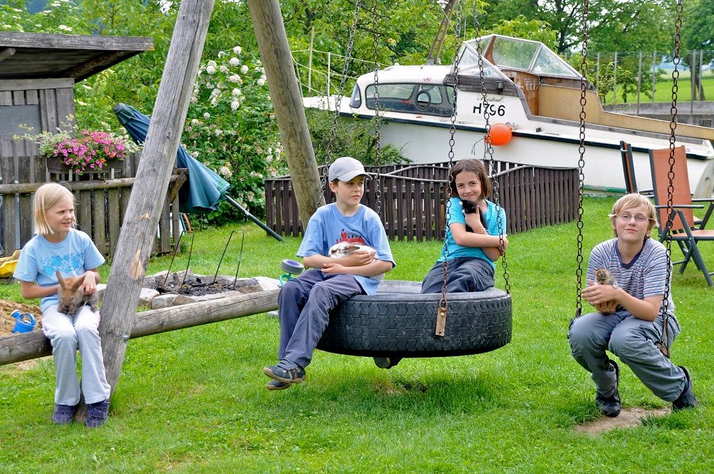 Four children sit on a swing in the garden and hold animals. There is a boat in the background.