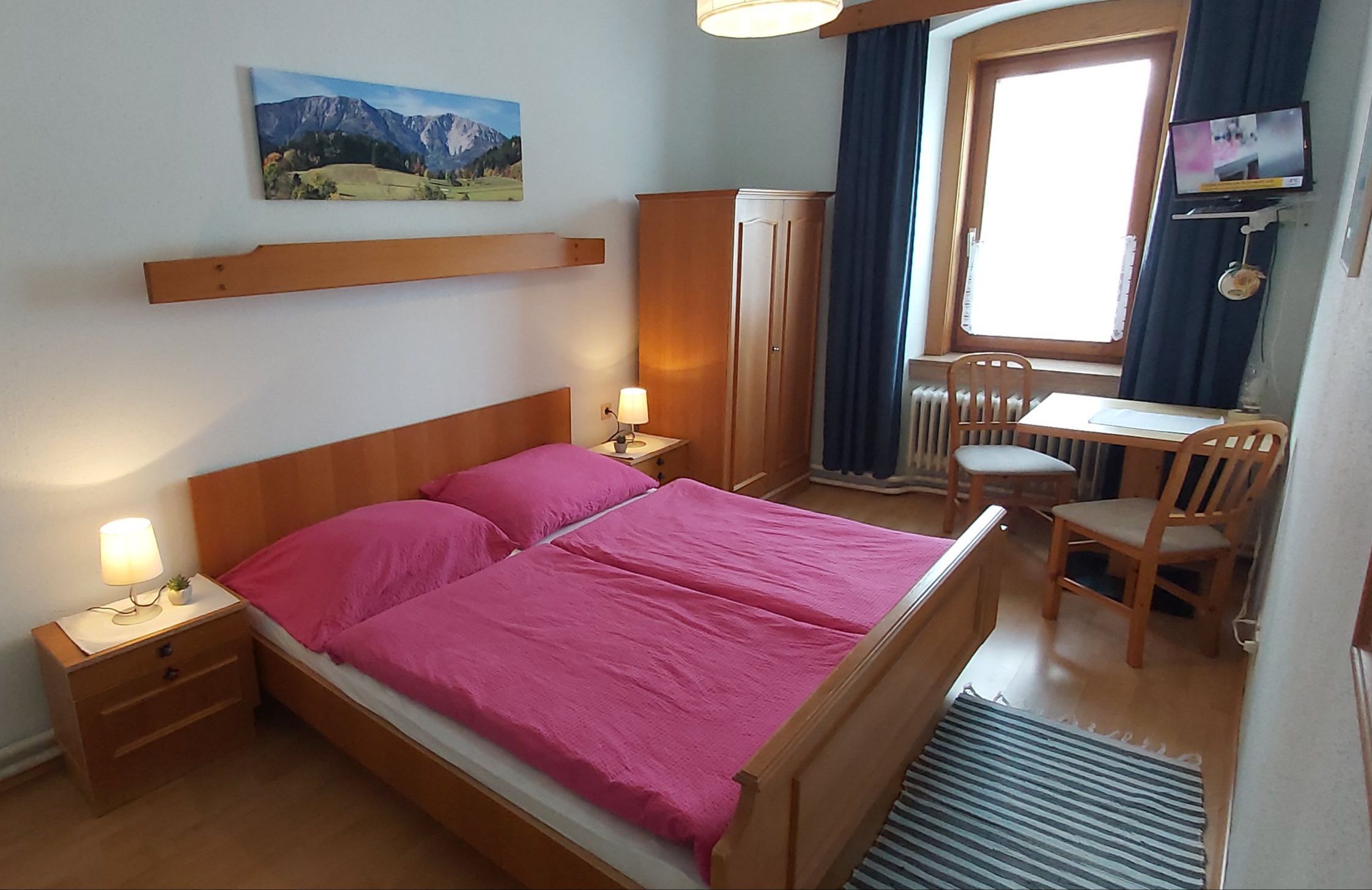 A cozy double room with wooden furniture, a double bed with pink bed linen, a table with two chairs and a small TV.