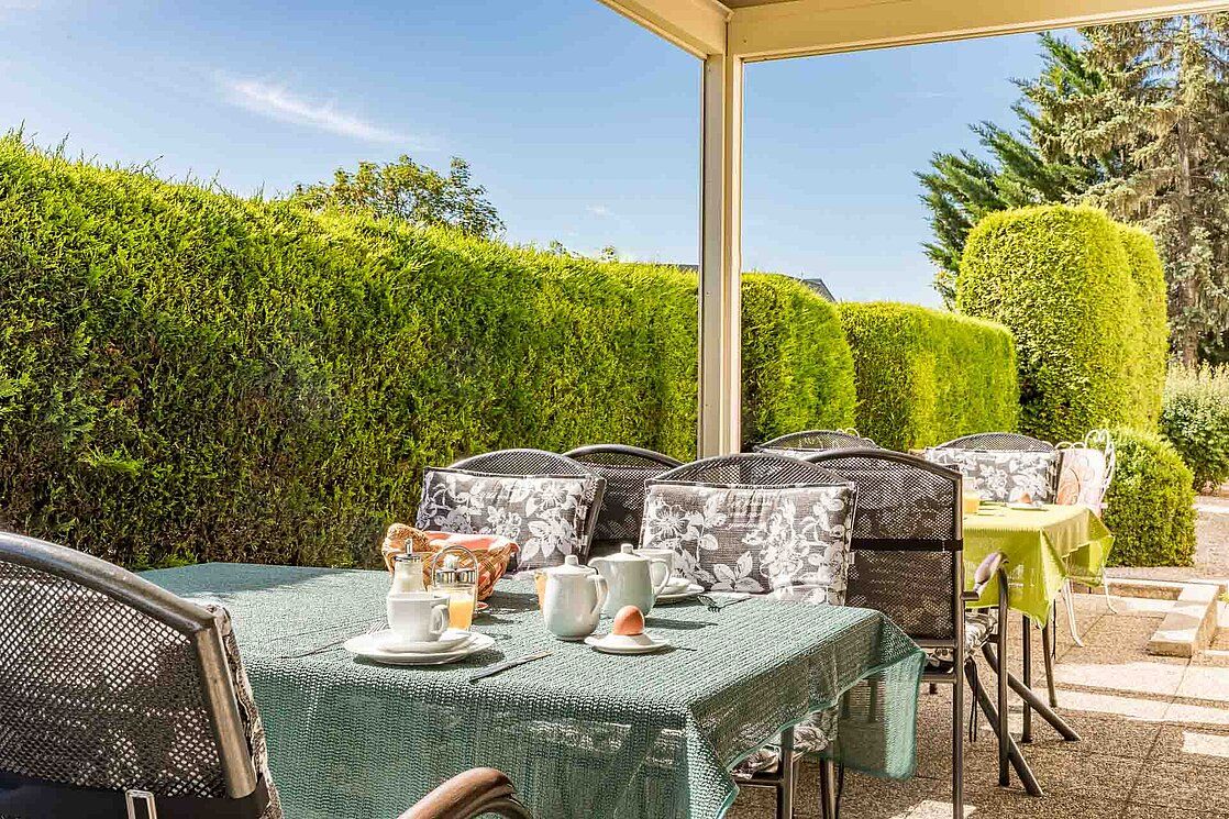 Outdoor breakfast table with crockery and green tablecloth, surrounded by hedges.
