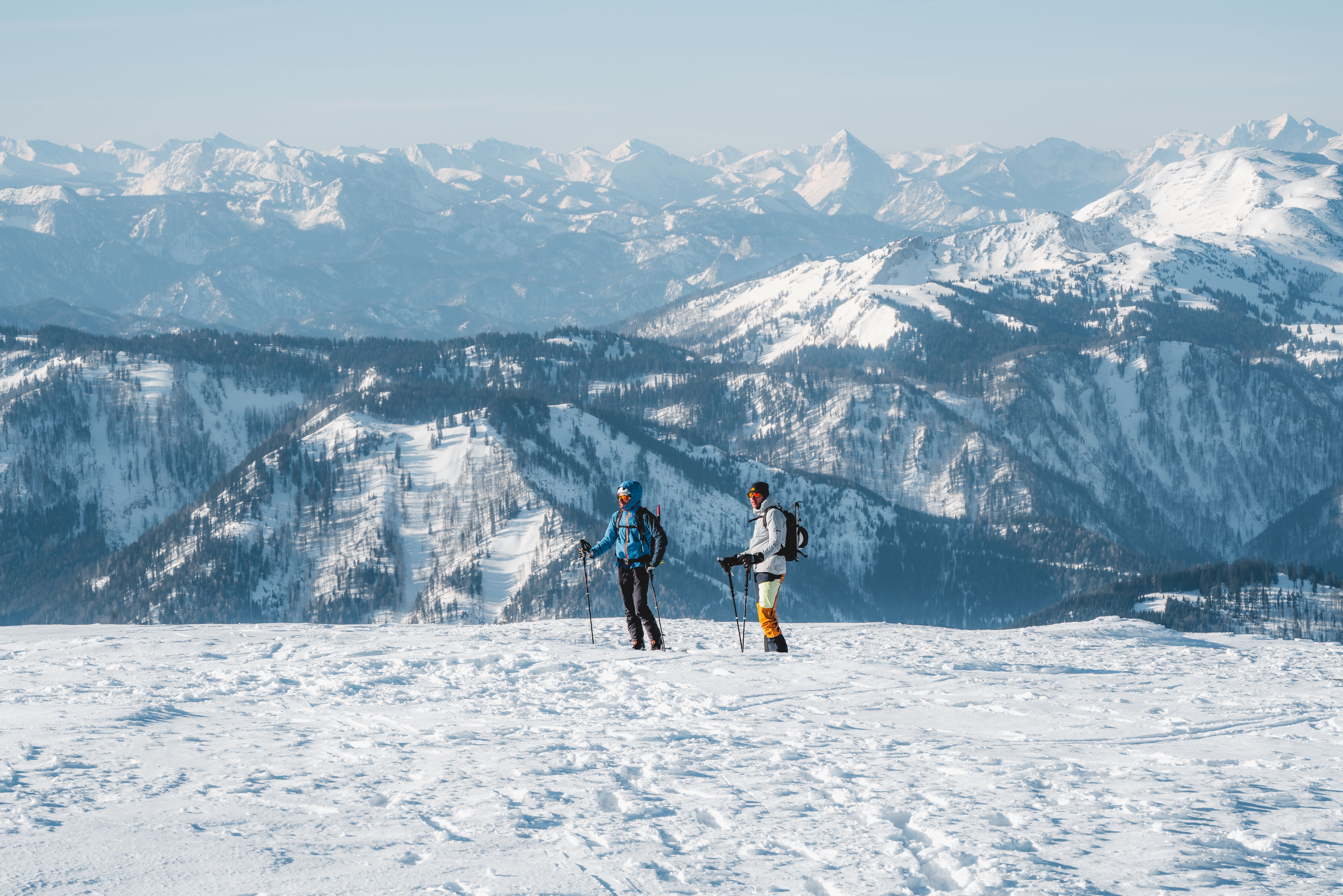 Die glitzernde Schneedecke erstreckt sich bis zum Horizont, während zwei Skitourengeher die unberührte Winterlandschaft erkunden. Die majestätischen Berge im Hintergrund bieten eine atemberaubende Kulisse und laden dazu ein, die Ruhe und Schönheit der Natur zu genießen.