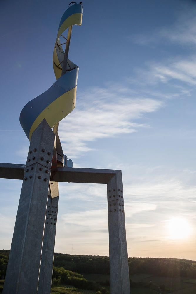 A large, spiral-shaped metal sculpture, reminiscent of a corkscrew, stands in front of a blue sky with a few clouds and the setting sun.