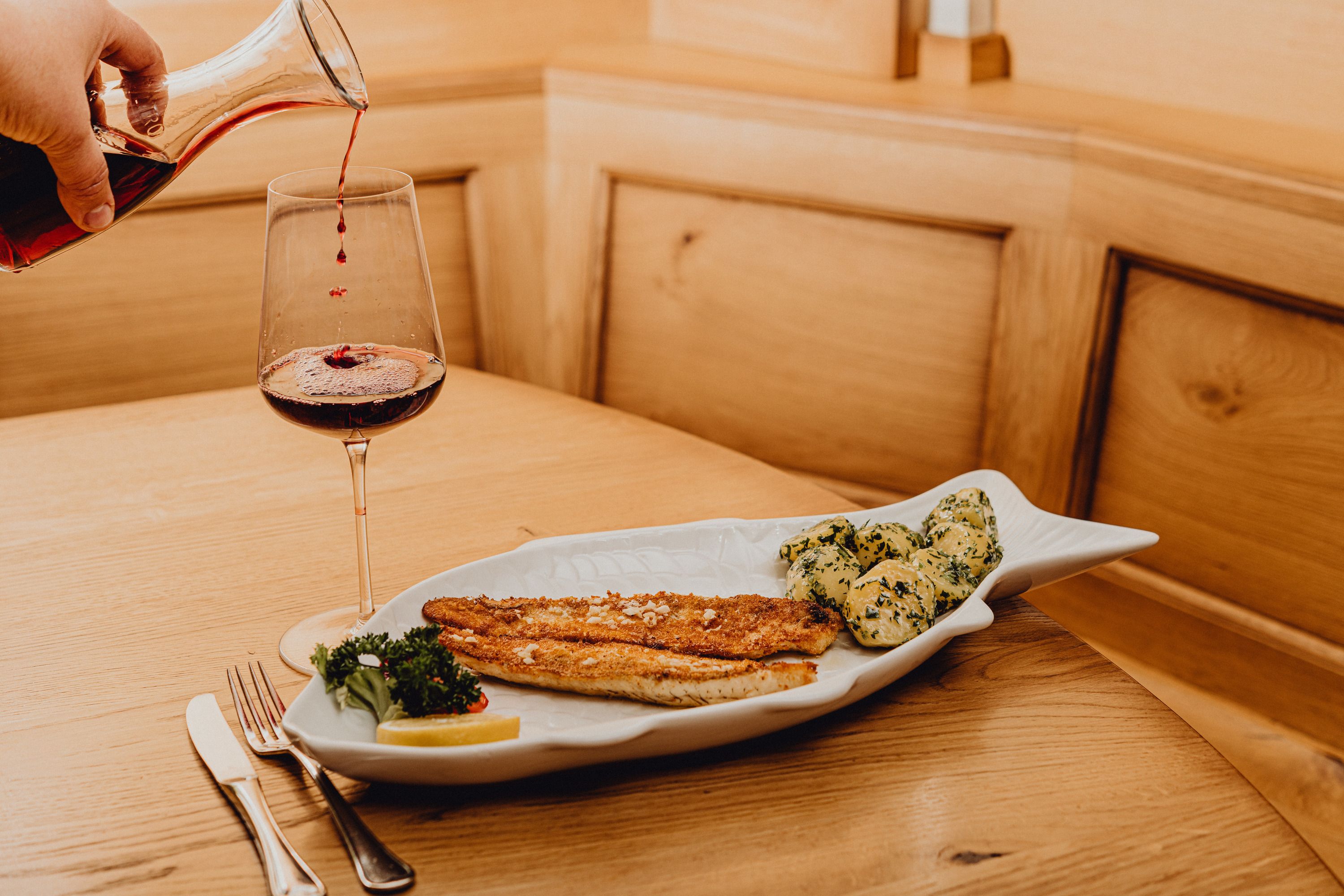 A plate with pike-perch fillet and potatoes in garlic butter, next to it a glass of red wine on a wooden table.