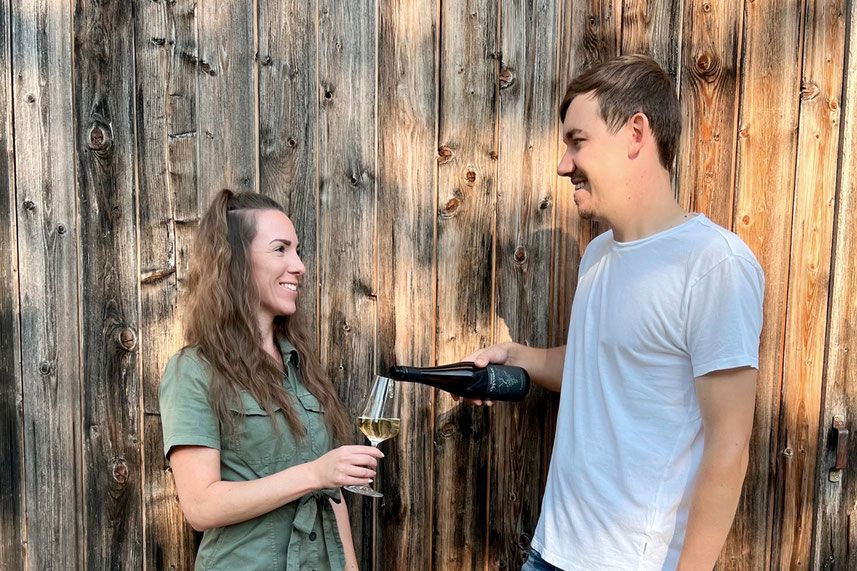 A man pours wine for a woman, both smiling in front of a wooden wall.