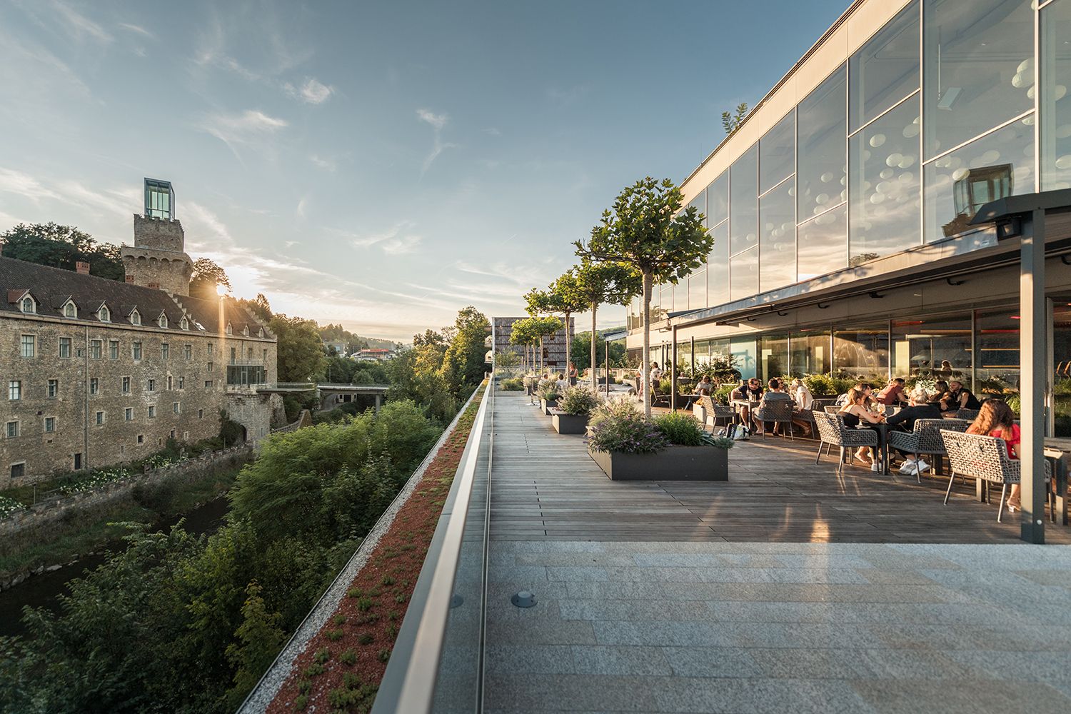 Panorama of a river landscape with a castle and modern architecture, surrounded by green hills.