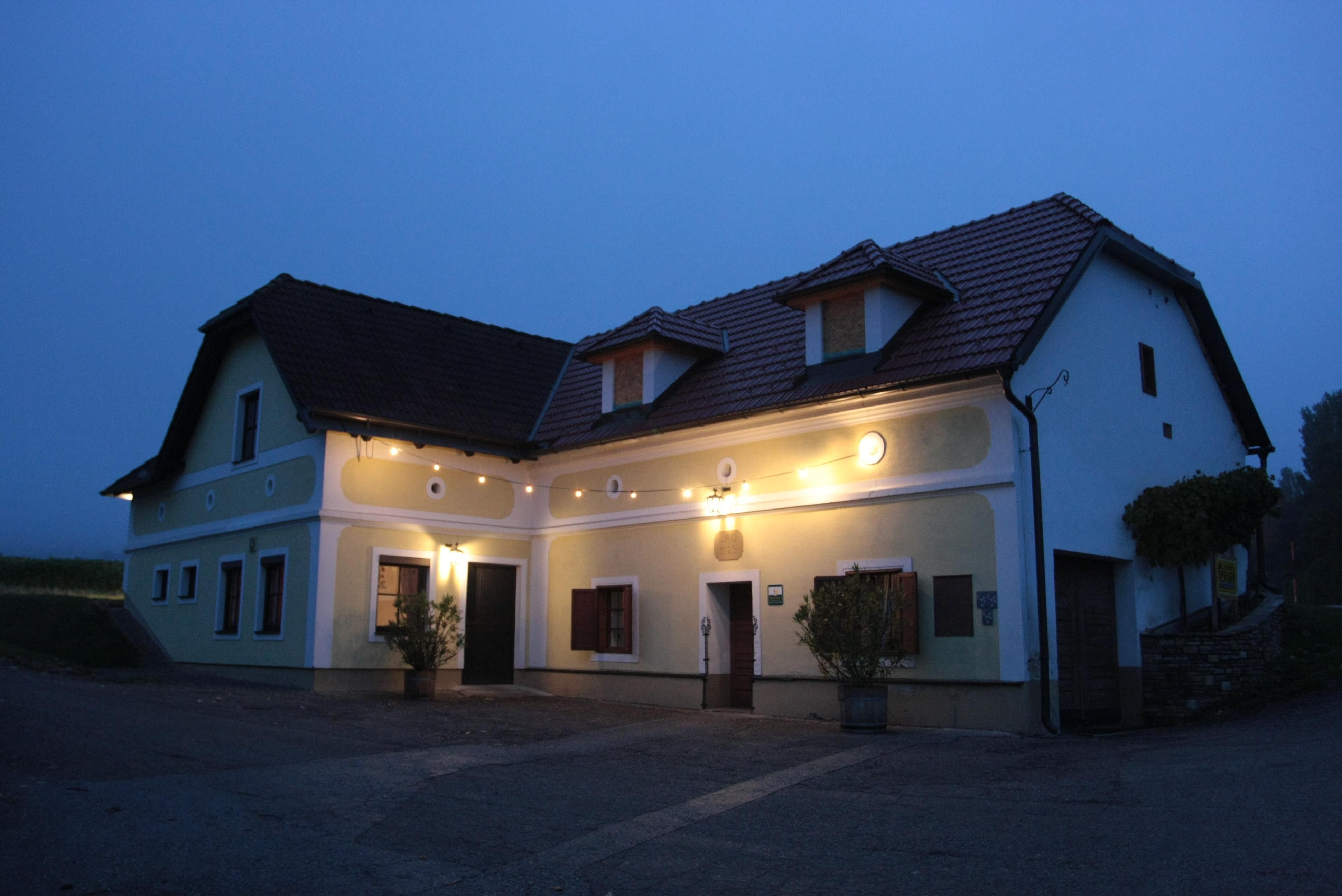 A traditional, illuminated house with a yellow façade and red roof tiles at dusk.