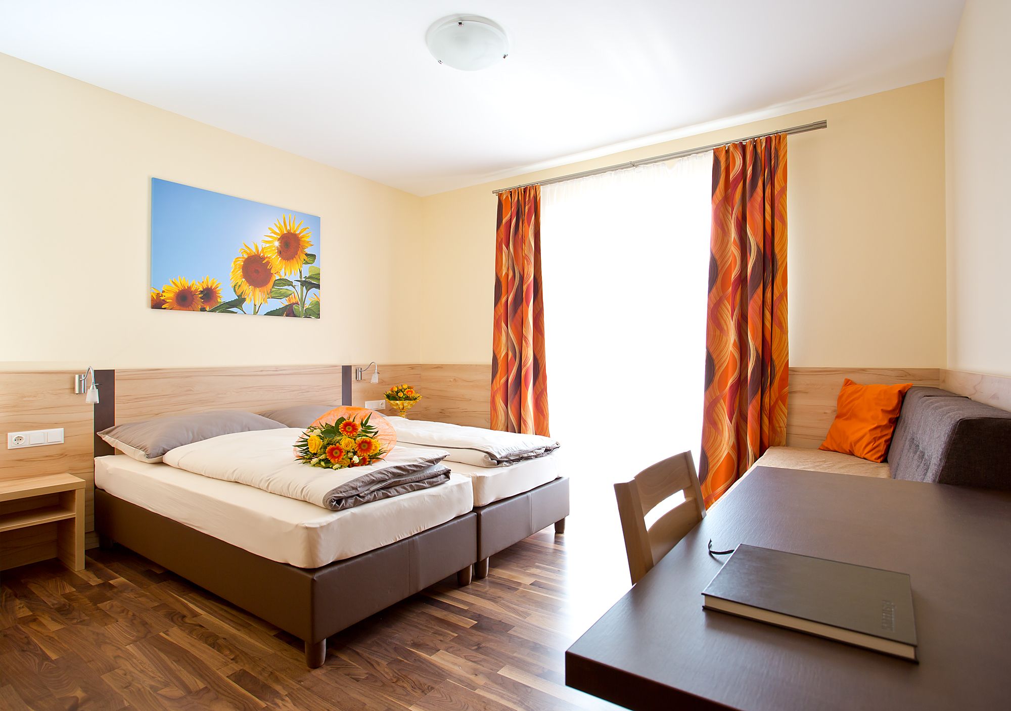 A bright guest room with two beds, a table, a sofa and a picture of sunflowers on the wall.
