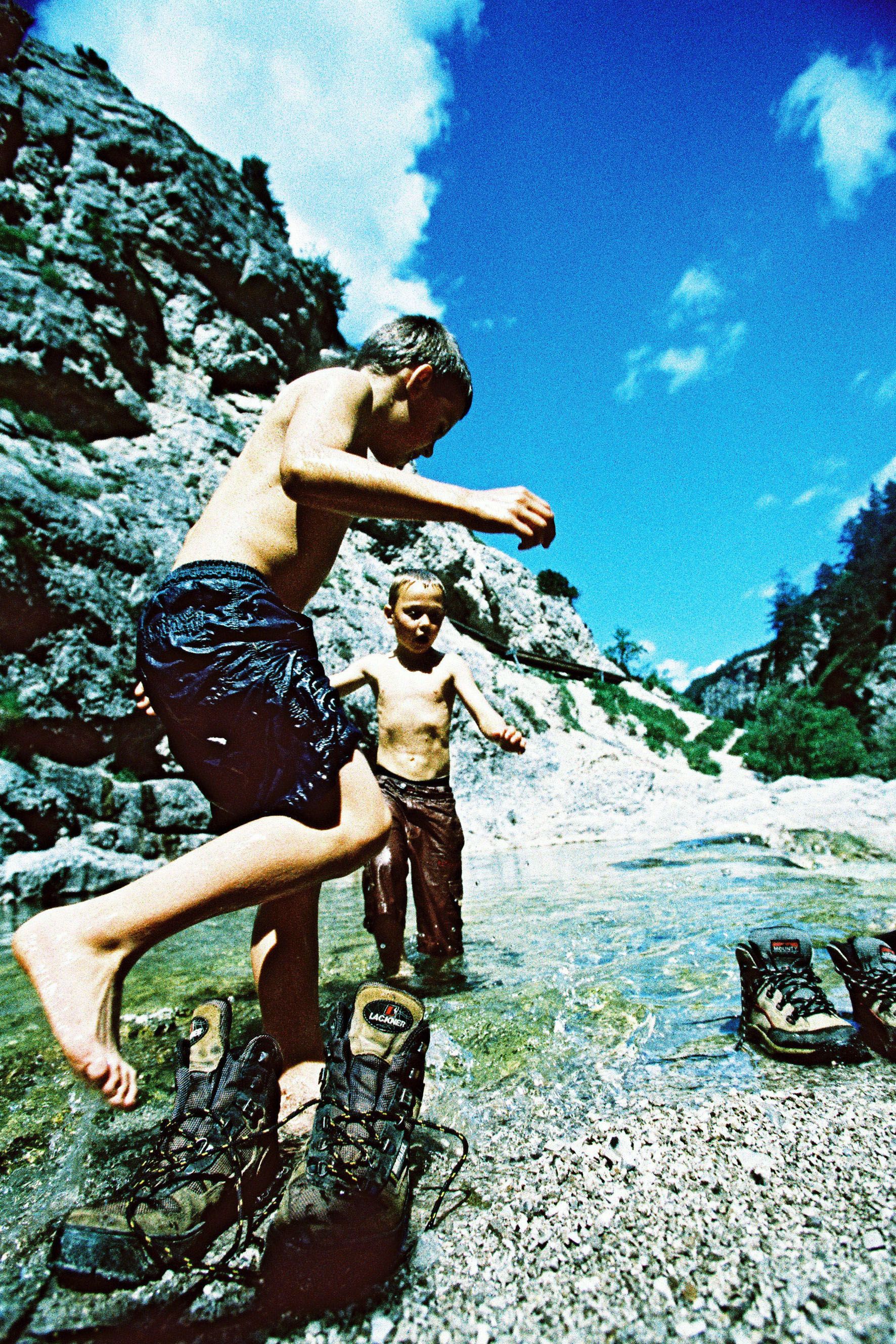Two children play in the water of a mountain stream, surrounded by rocks and blue sky.