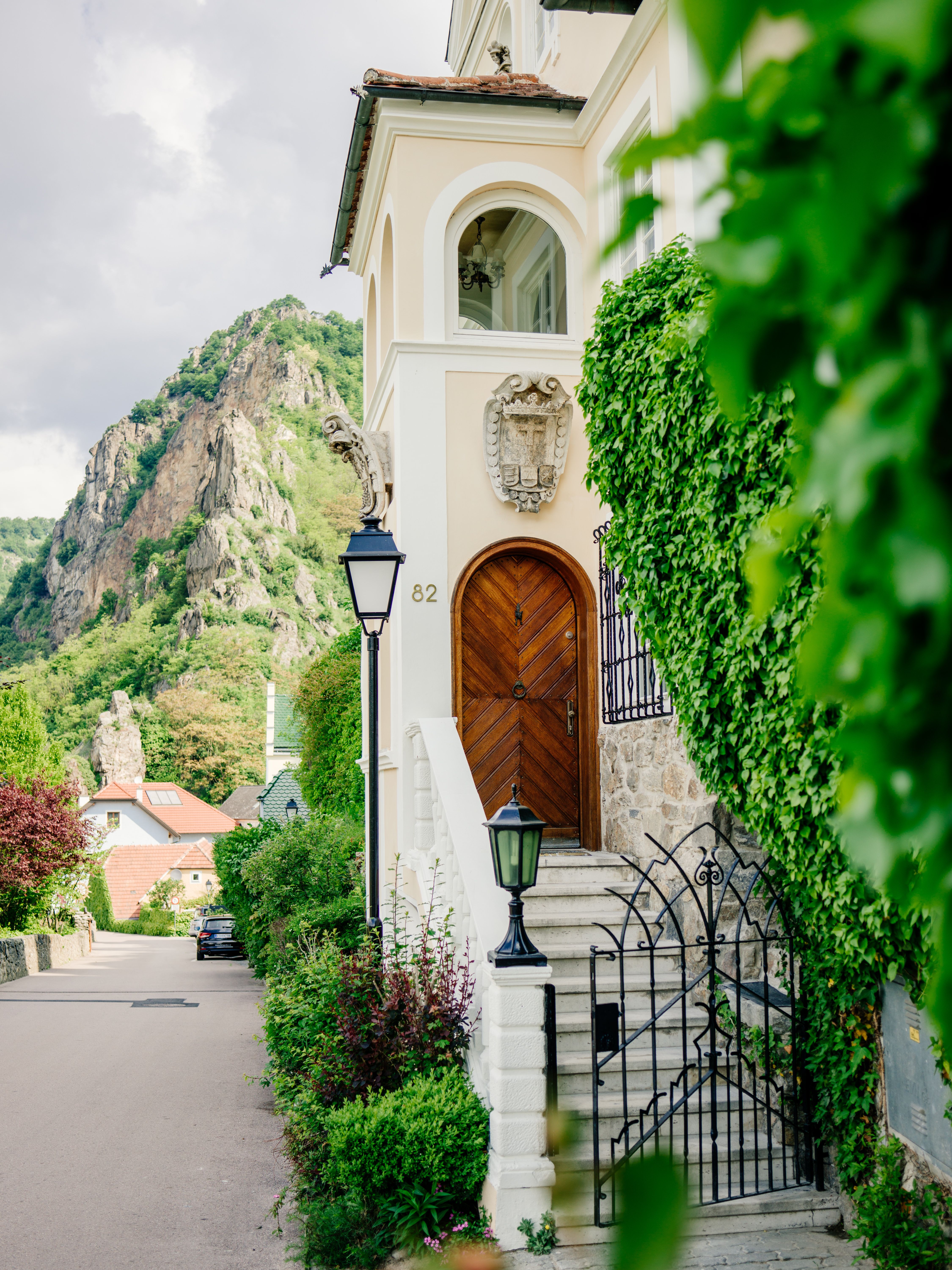 Entrance to a villa with wooden door and lantern, surrounded by green plants, a rock in the background.