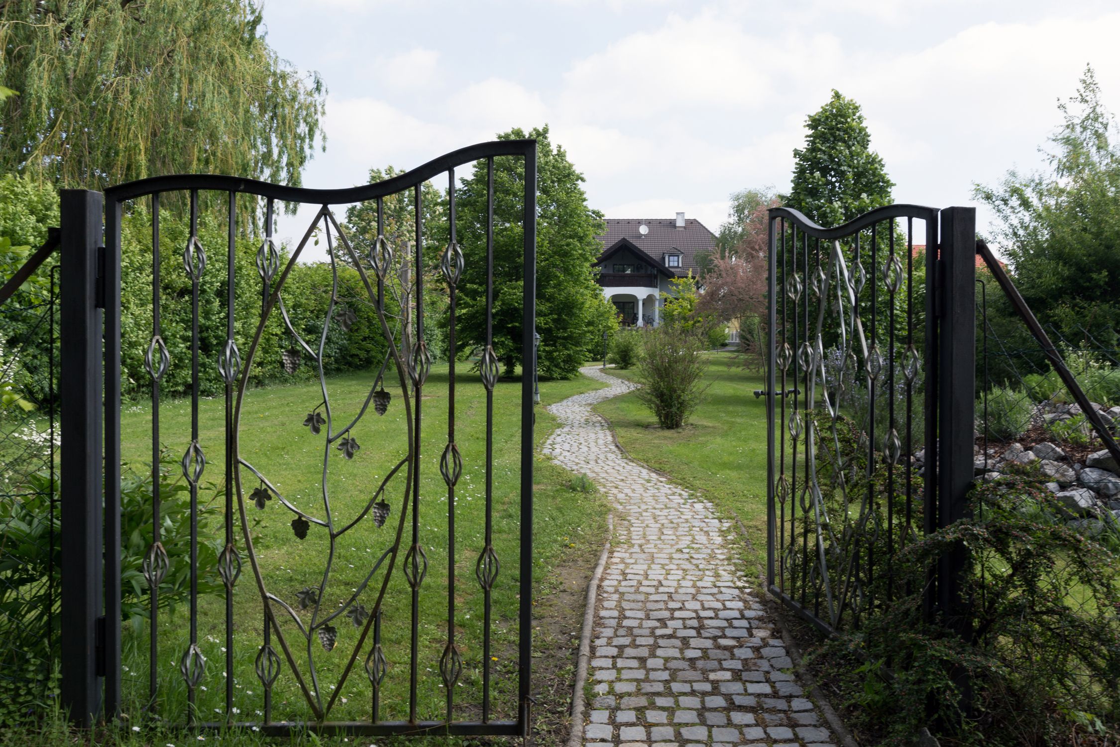 An open wrought-iron gate leads to a paved path that leads through a green garden to a house in the background.