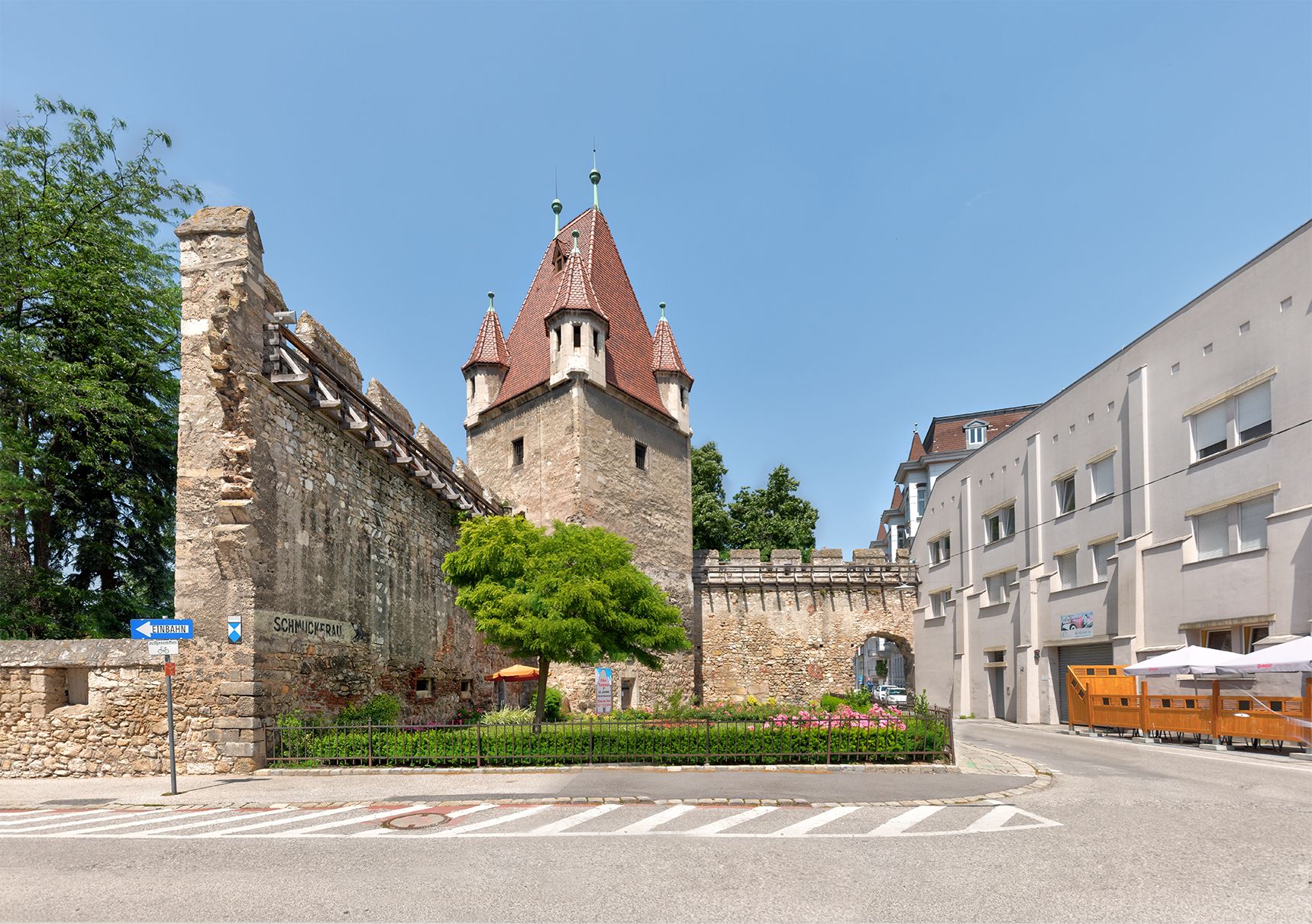 Historic stretching tower with red roof and adjoining wall, surrounded by modern buildings and a road.