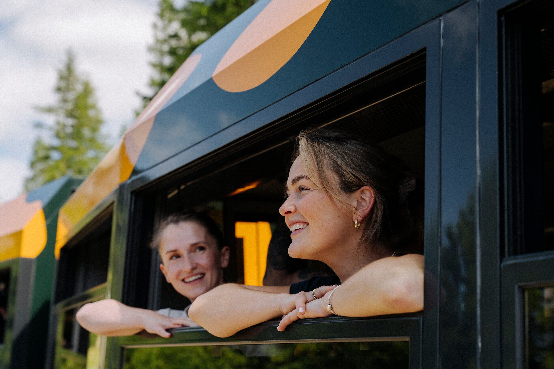 Two cheerful women look out the window of the Schneebergbahn's Salamander train as the wooded landscape passes by in the background, reflecting their joy and curiosity about the journey ahead.