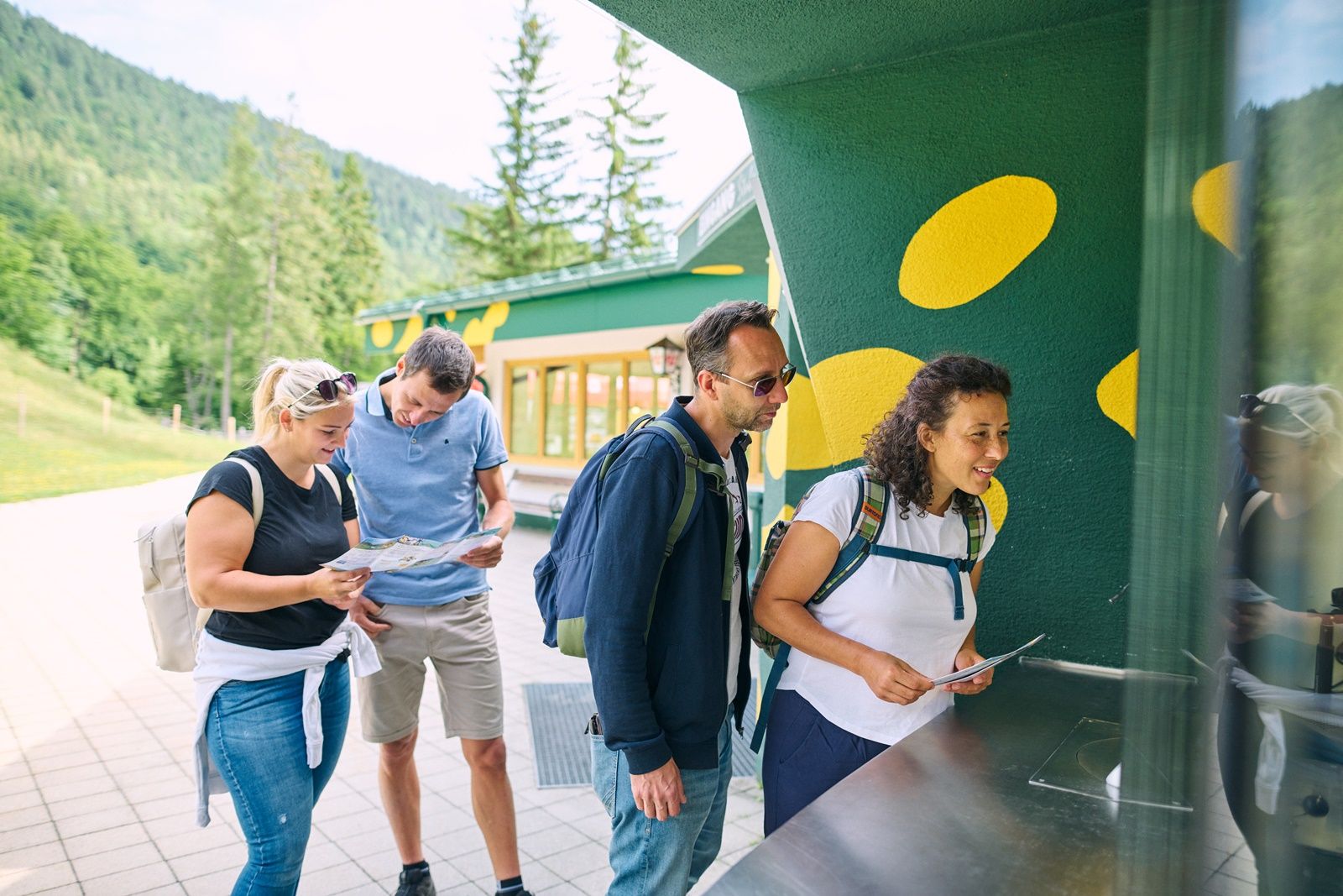 Four people stand at an outdoor ticket counter, surrounded by a green landscape.