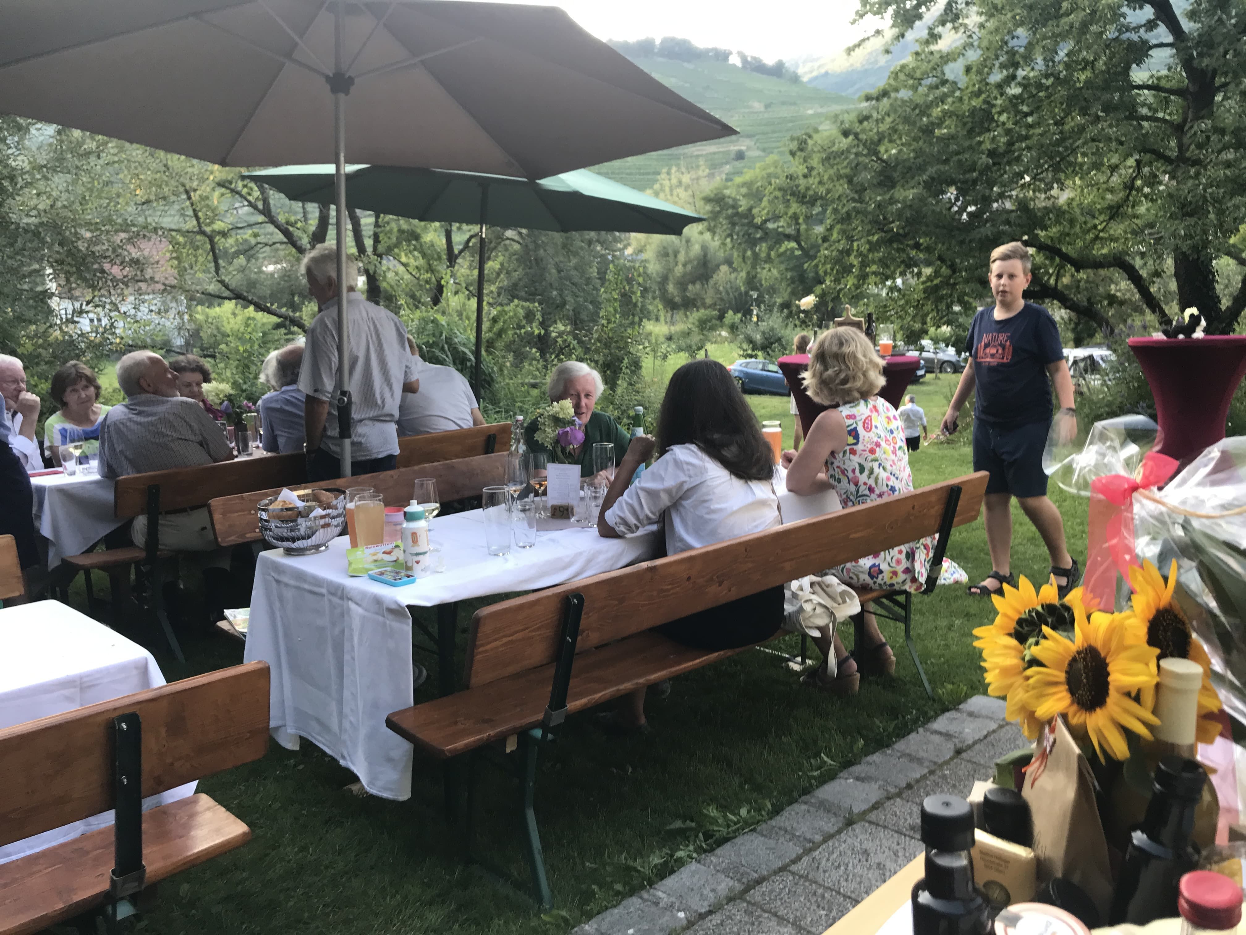 People sit outside at wine tables under parasols, surrounded by trees and meadows.