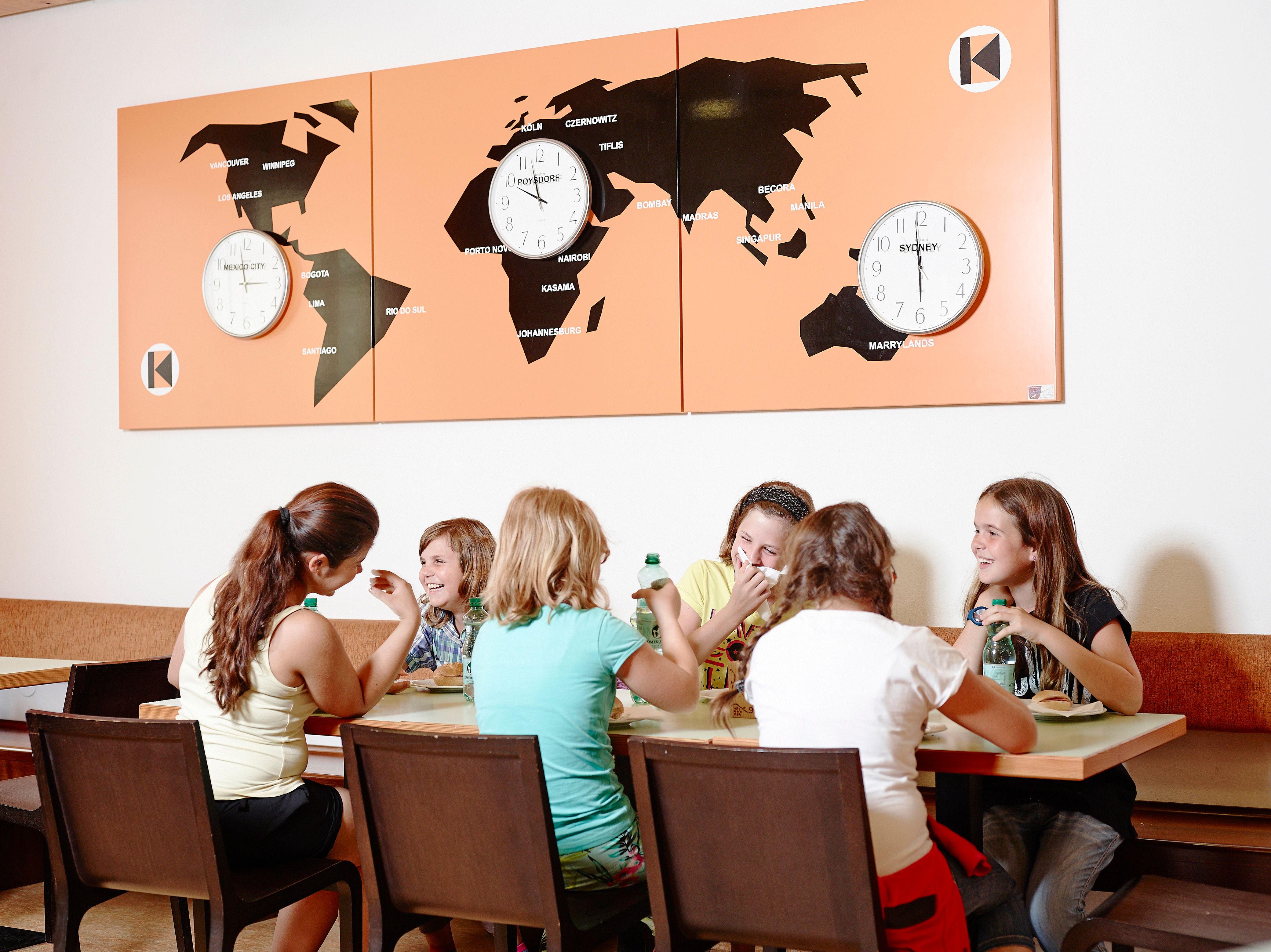 Young people sit at a table and eat, world maps with clocks in the background.