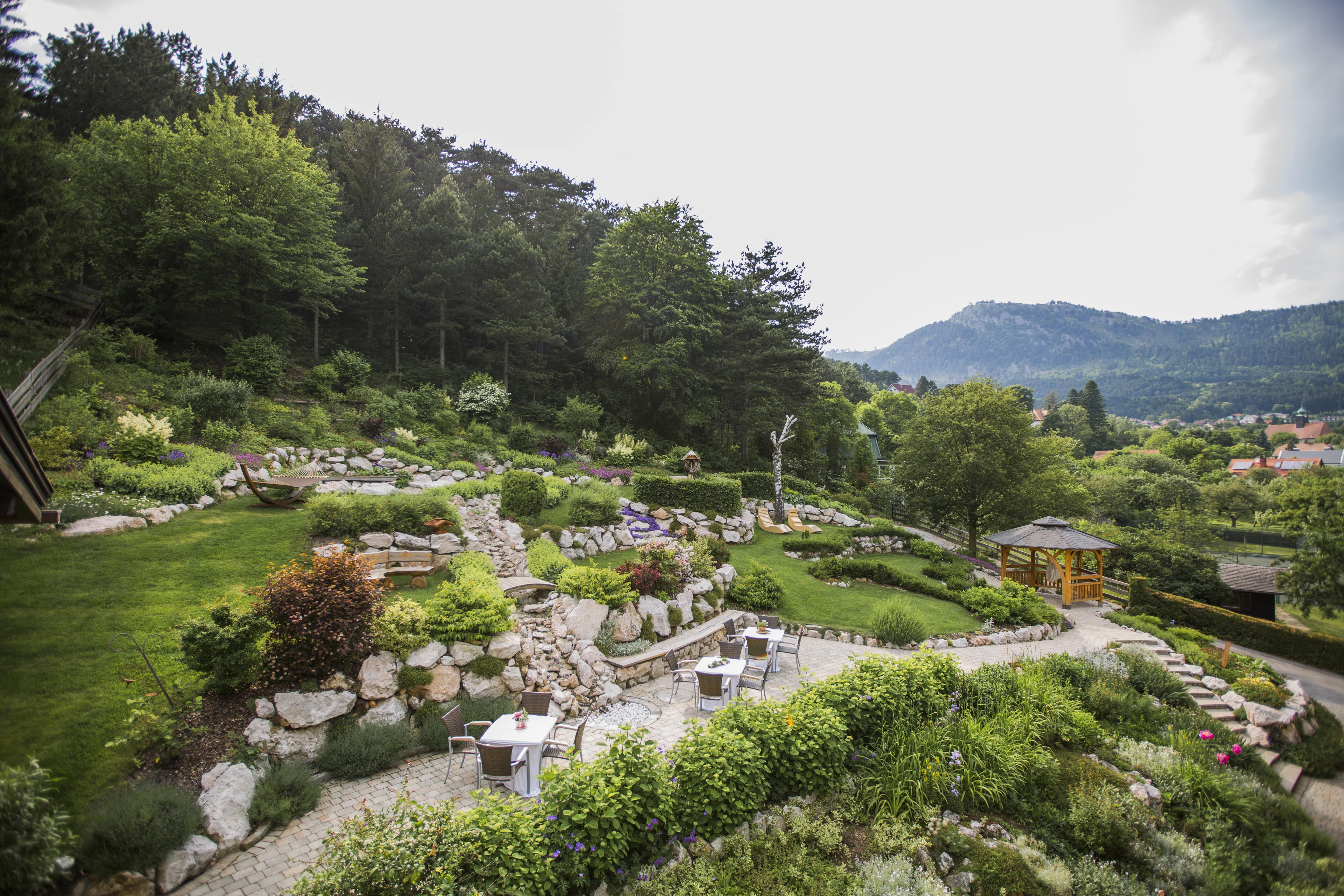 A terraced garden with stones, plants and seating areas, surrounded by trees and mountains in the background.