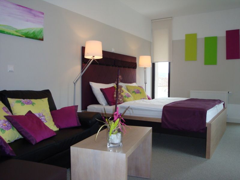 Modernly furnished hotel room with double bed, sofa and decoration in shades of purple and green.