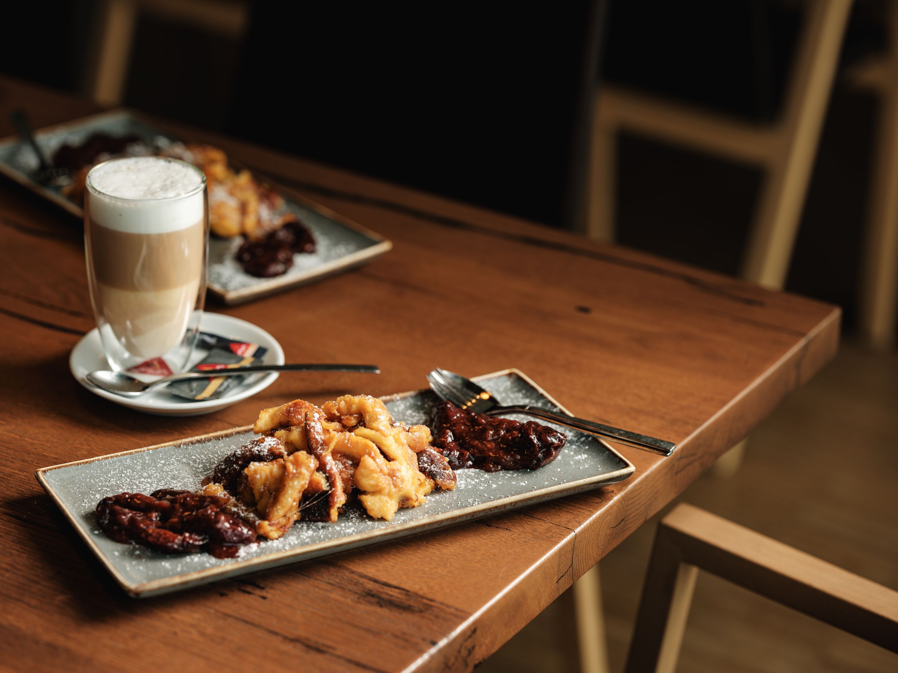 A plate of Kaiserschmarrn and stewed plums on a wooden table with a glass of latte macchiato next to it.