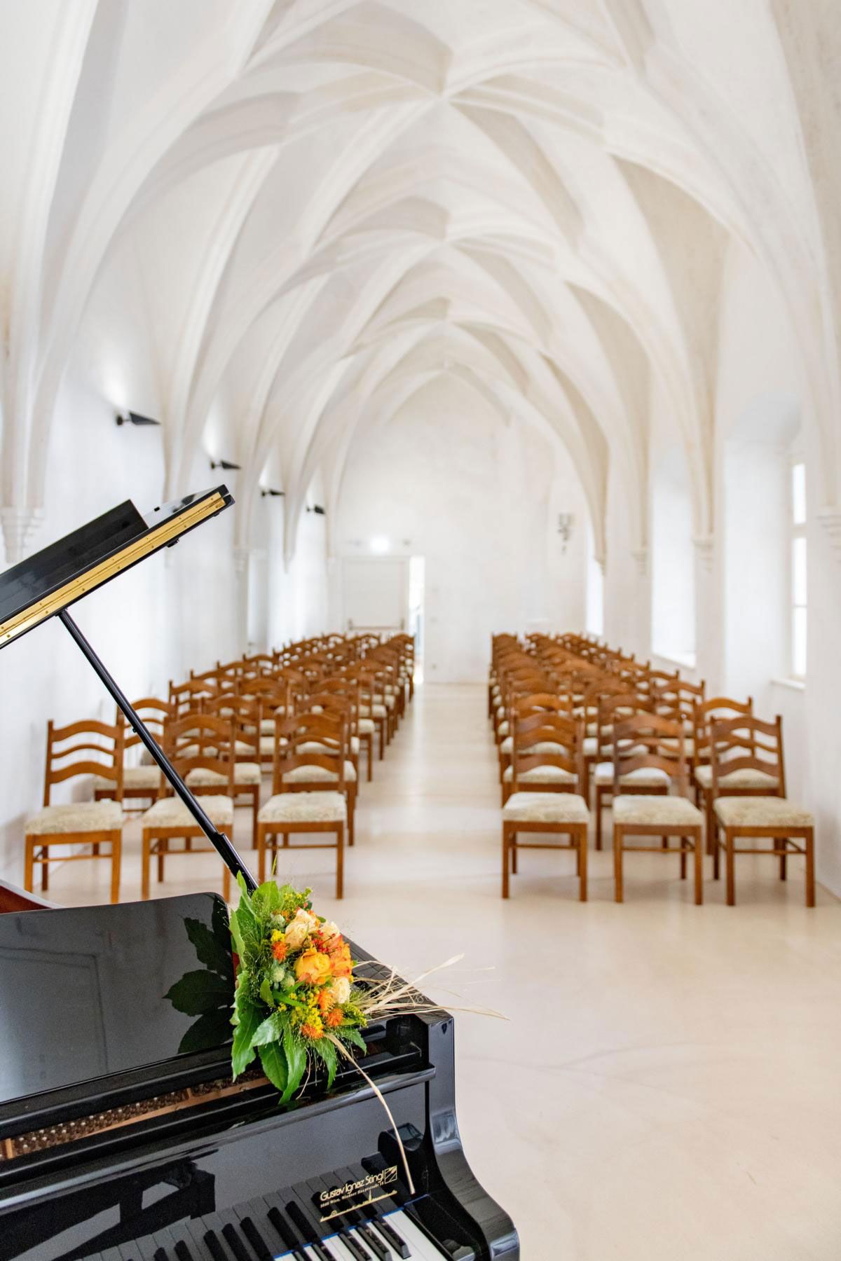 A grand piano with flower arrangements stands in a Gothic hall with a vaulted ceiling and rows of wooden chairs.