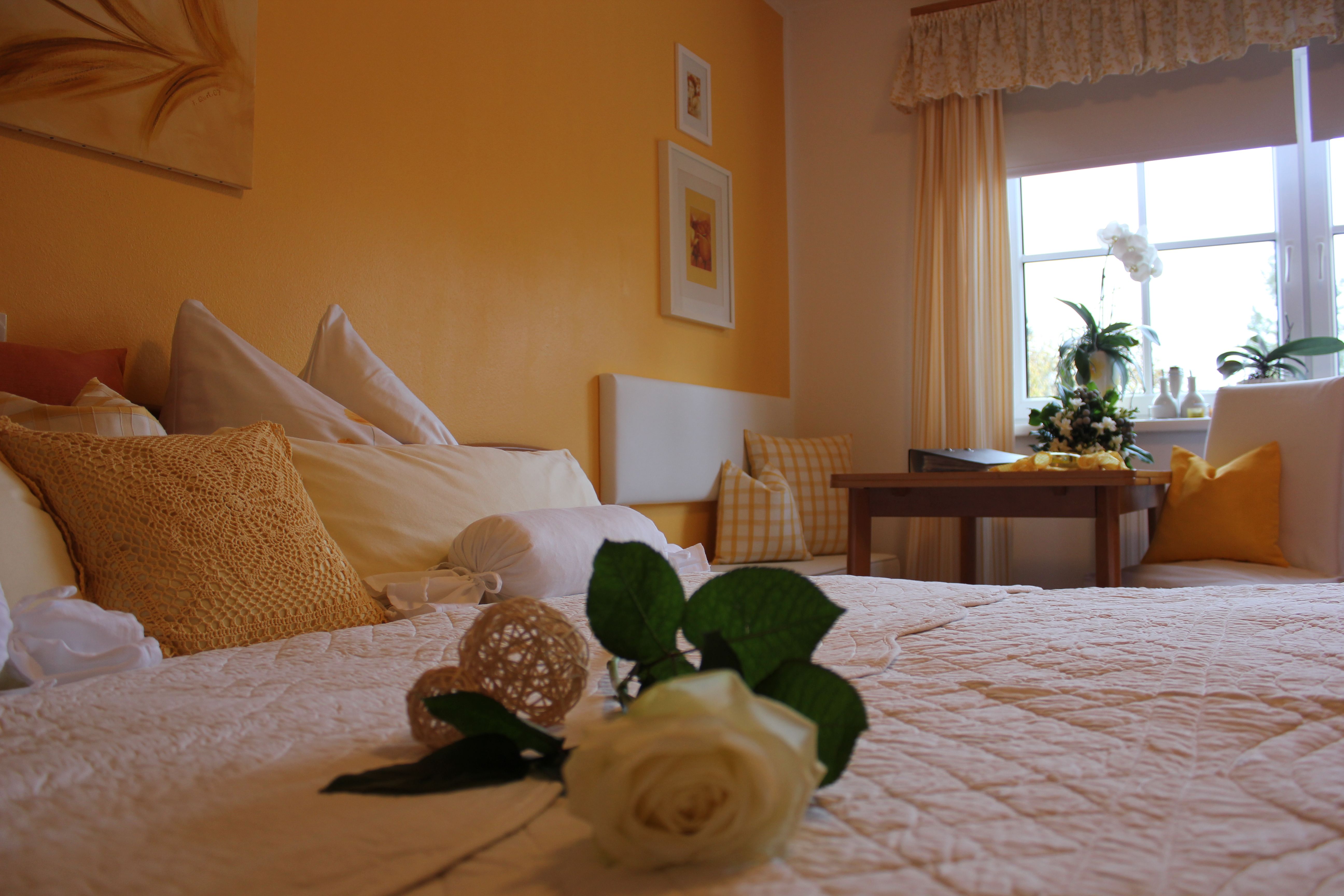 Cozy bedroom with yellow walls, a bed with white and yellow pillows, a table and a window with plants.