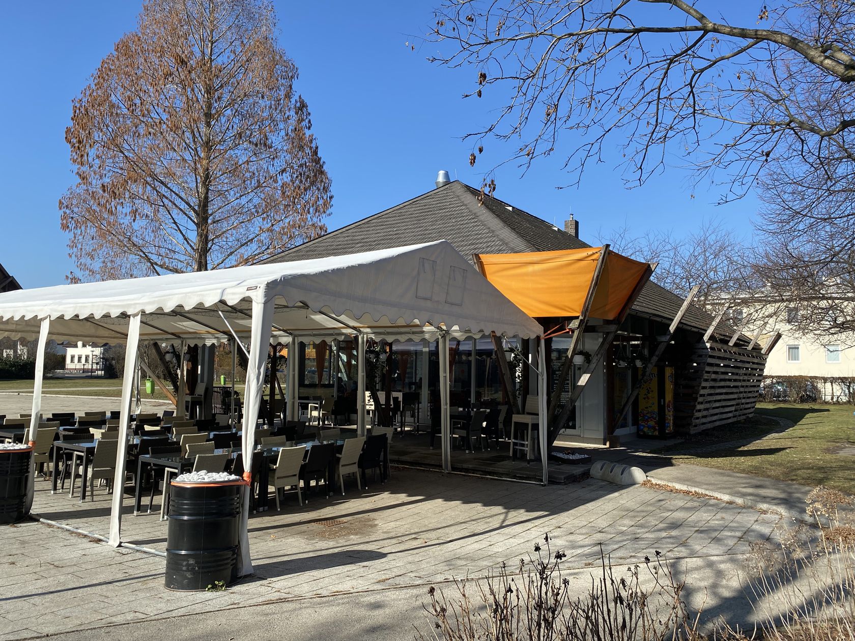 A café with an outdoor seating area in Eduard Garderer Park, surrounded by bare trees and blue skies.