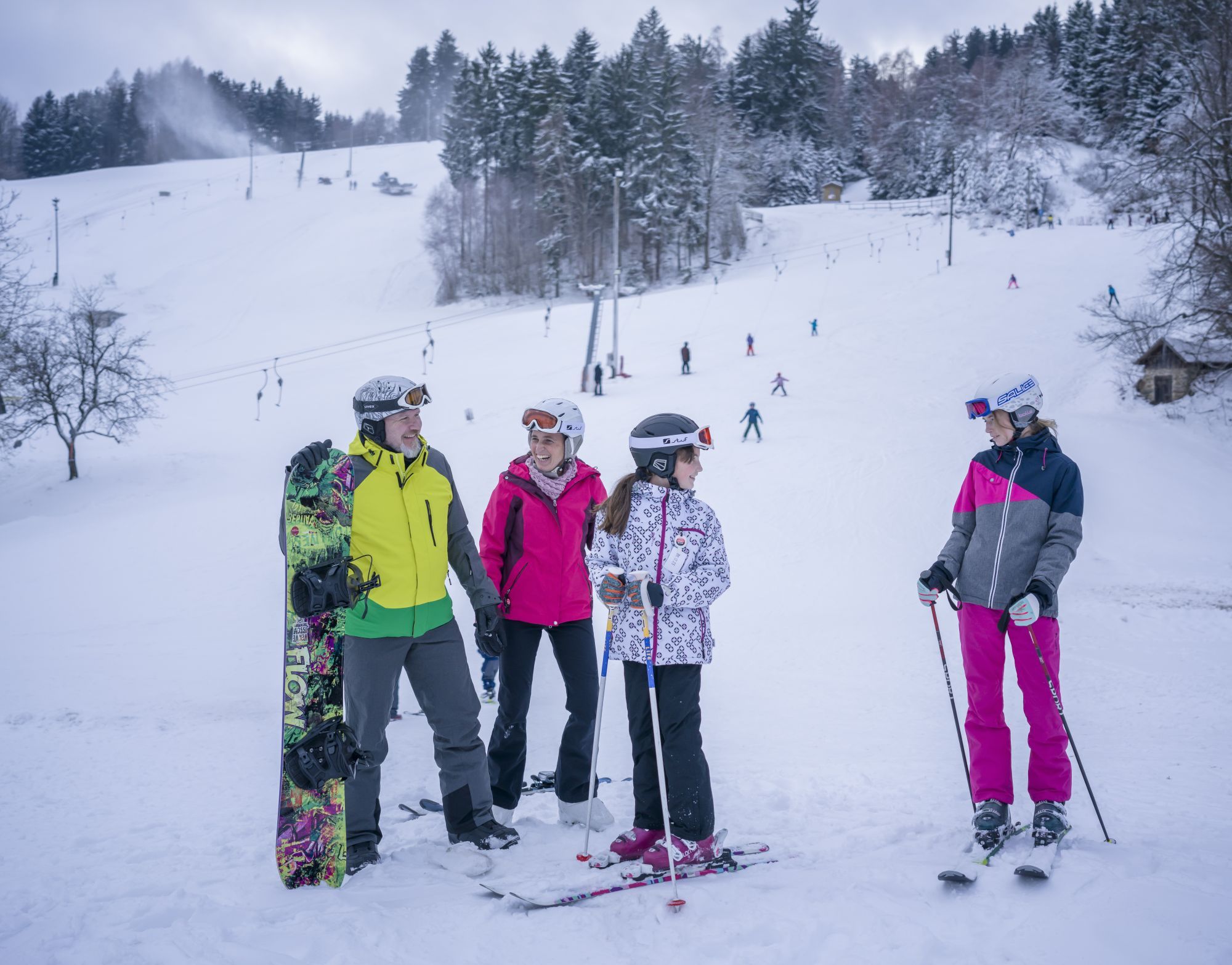 Group of skiers and snowboarders in the snow in front of a ski slope with a lift.