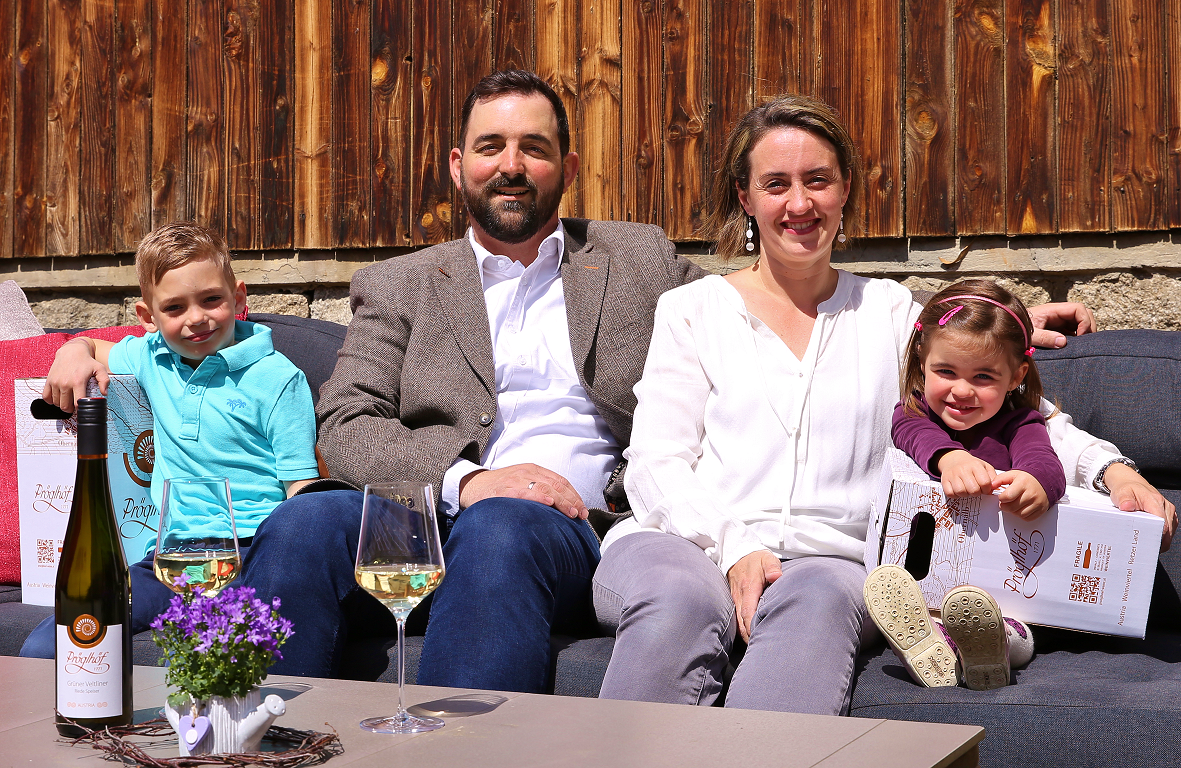 A family sits on an outdoor sofa in front of a wooden wall, with bottles of wine and glasses on the table.
