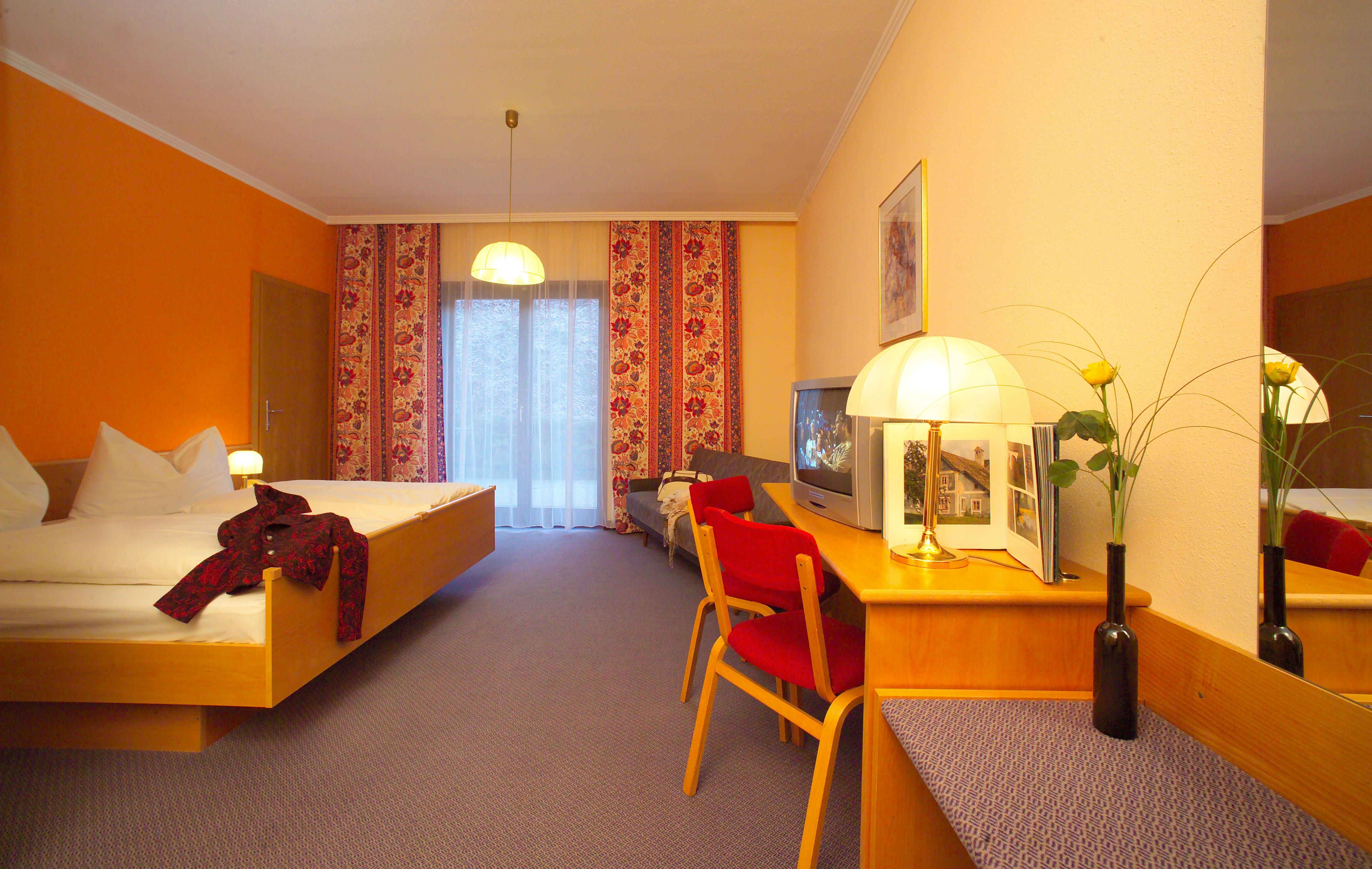 Hotel room with double bed, red chair, TV and lamp on a table. Yellow wall and curtains with a floral pattern.