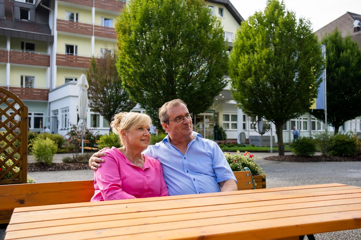 A couple sits on a bench in front of a building with trees in the background.