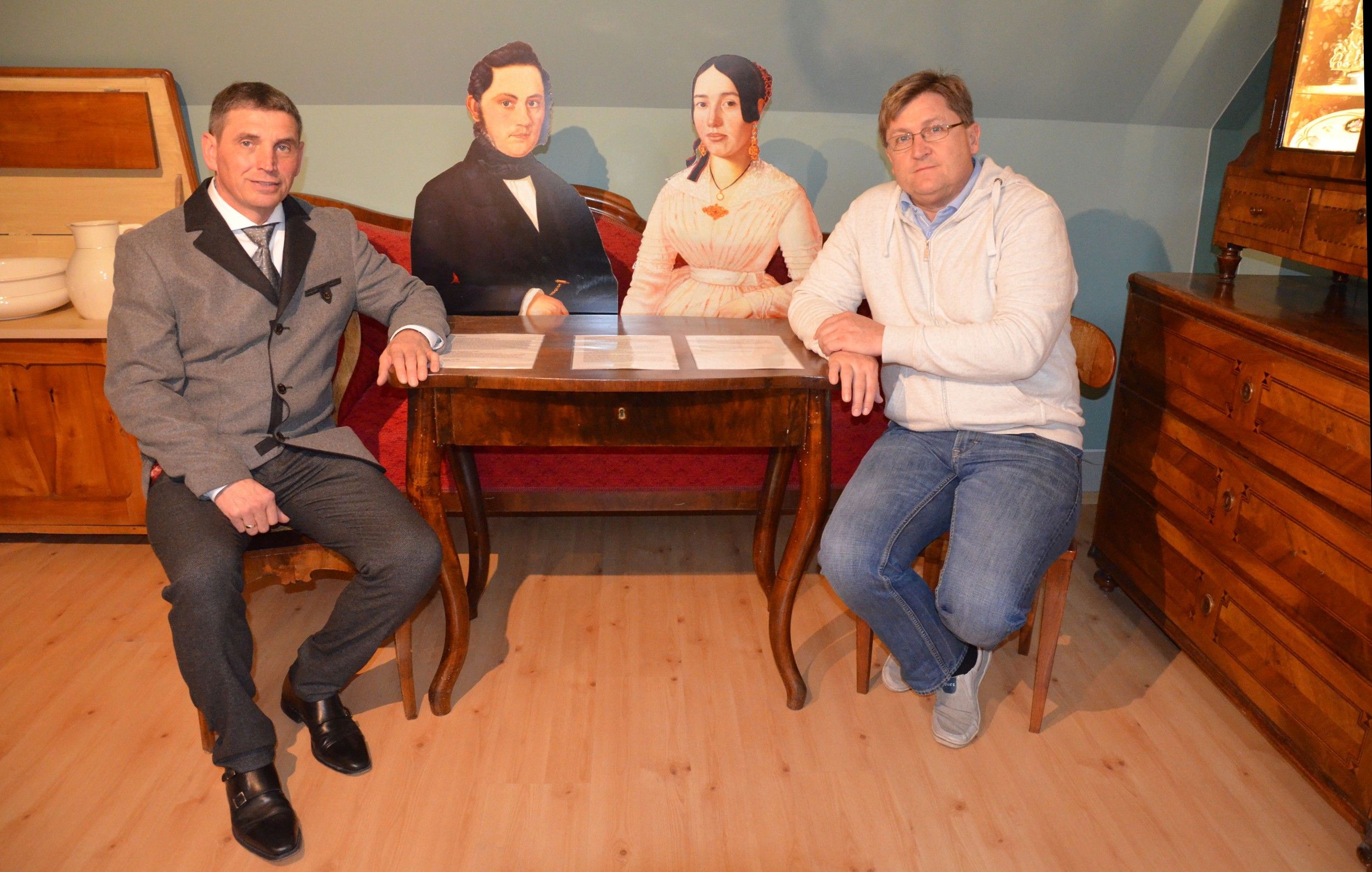 Two men sit at a table with two cardboard figures in a room with antique furniture.