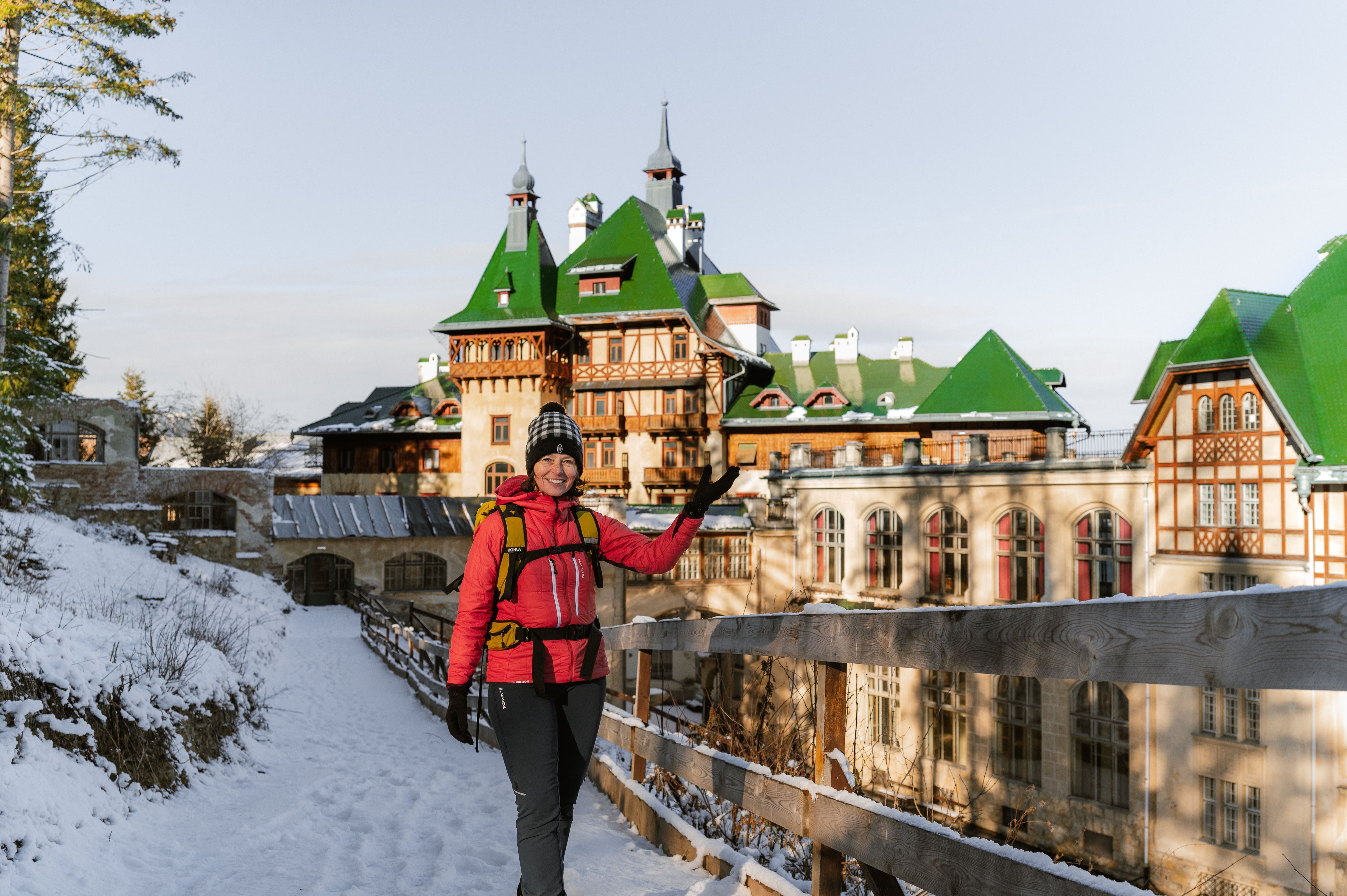 Person in winter clothing in front of a large building with green roofs in the snow.