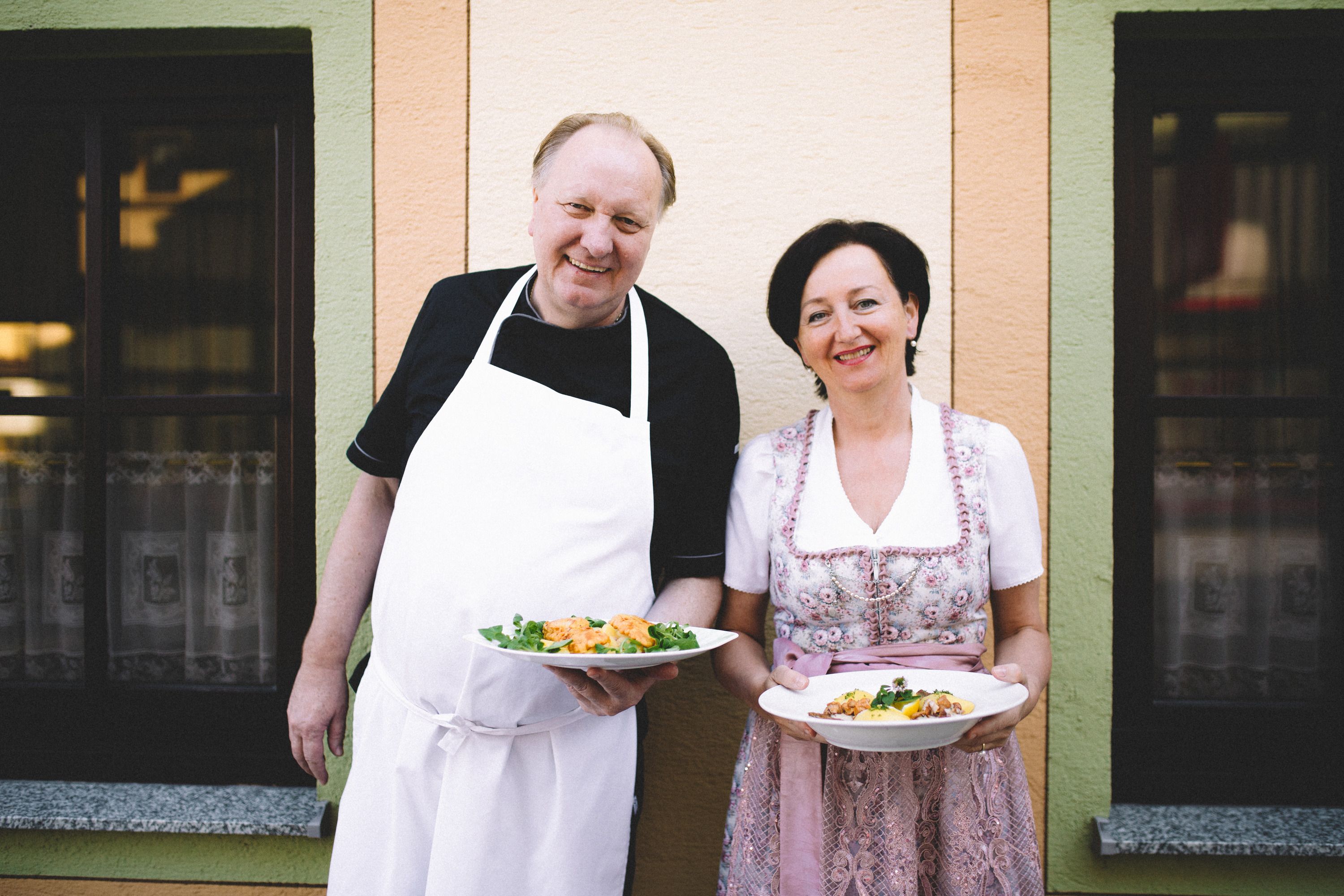 A man in an apron and a woman in traditional dress hold plates of food in front of a building.