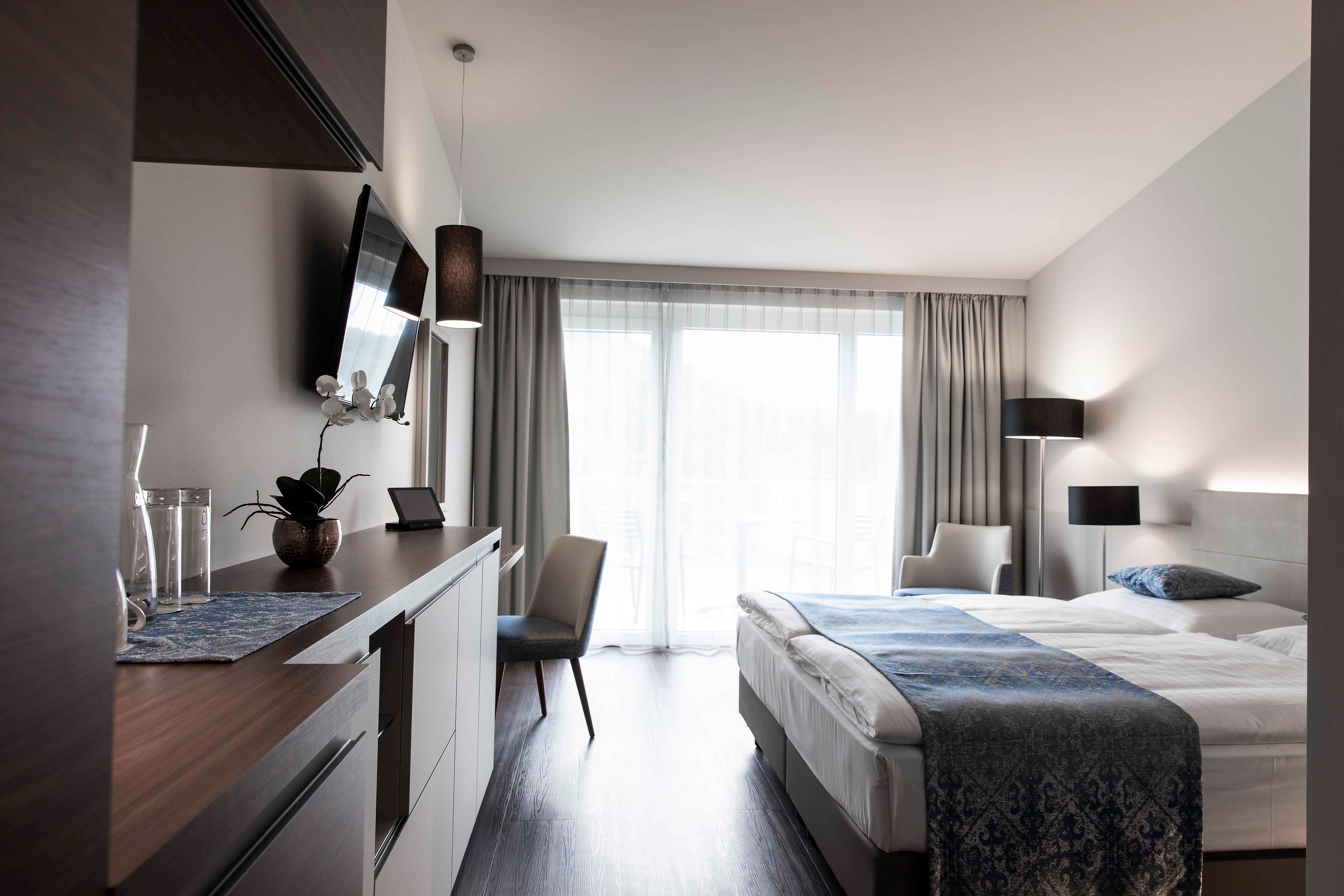 Modern hotel room with double bed, TV, desk and large windows with curtains.