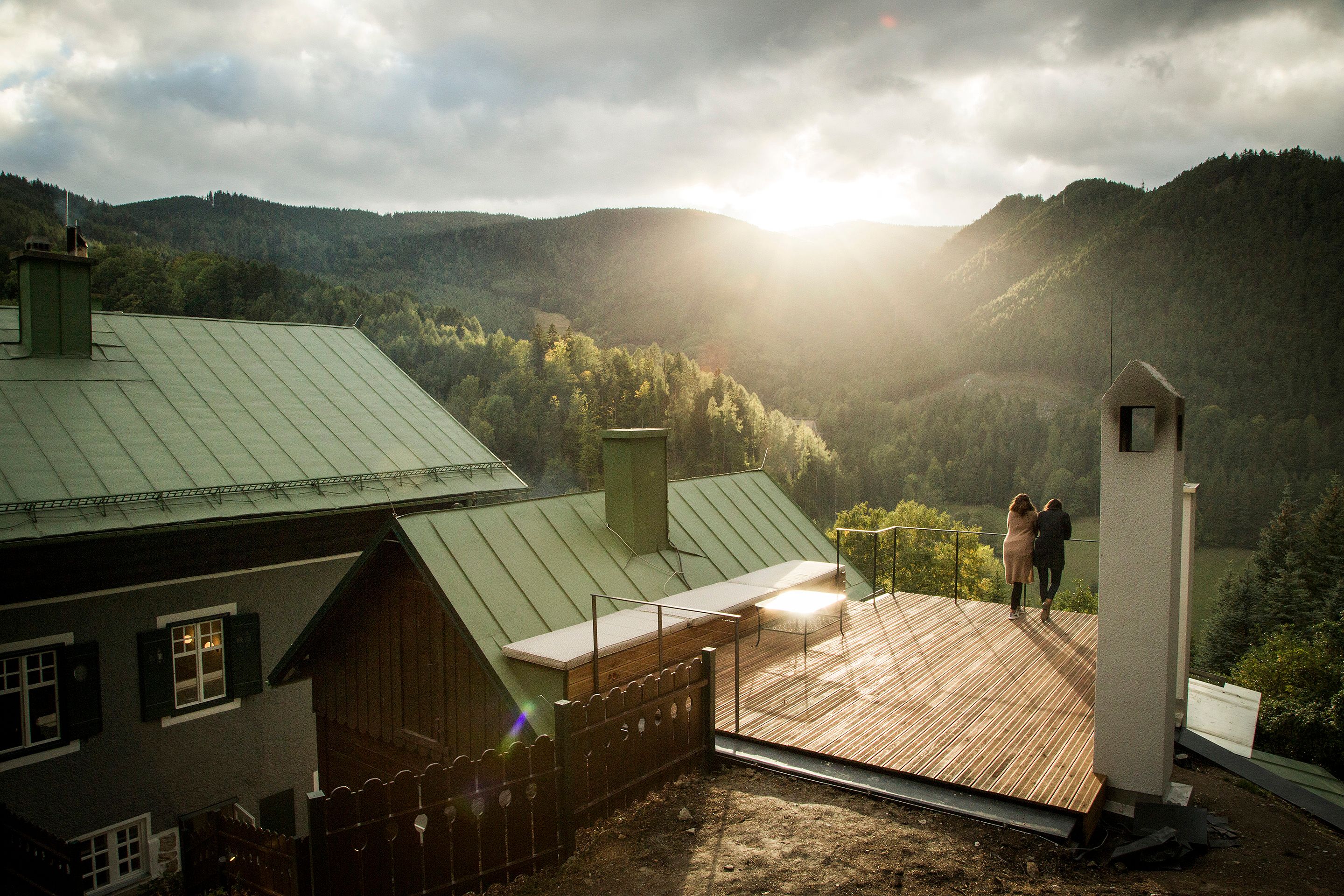 Two people stand on a terrace with a view of wooded mountains at sunset.