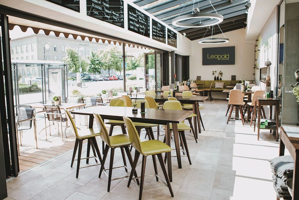 Modern interior of a wine bar with wooden tables and green chairs, large windows and a sign saying 'Leopold'.