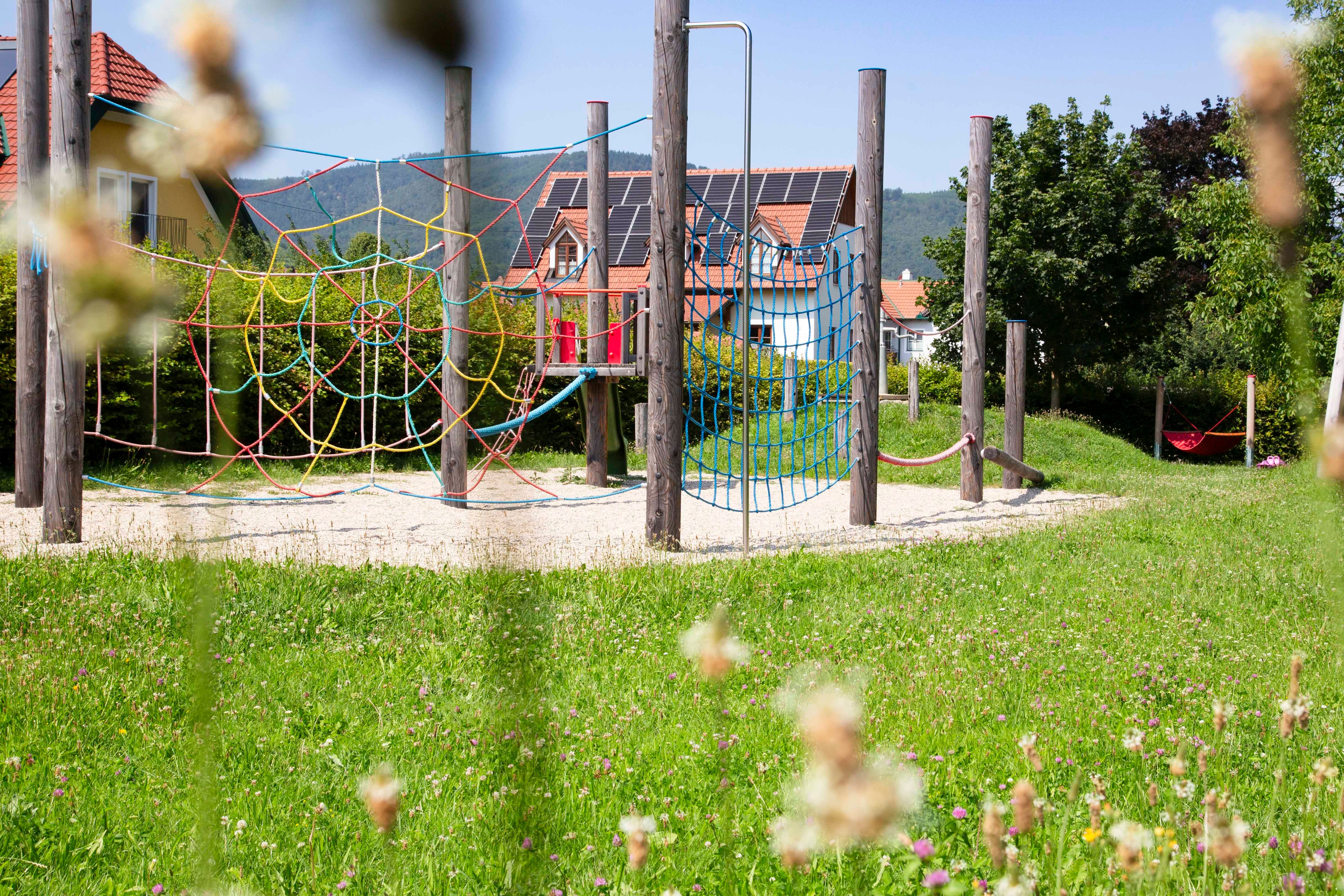 An adventure playground with climbing nets and wooden posts, surrounded by a green meadow and houses in the background.