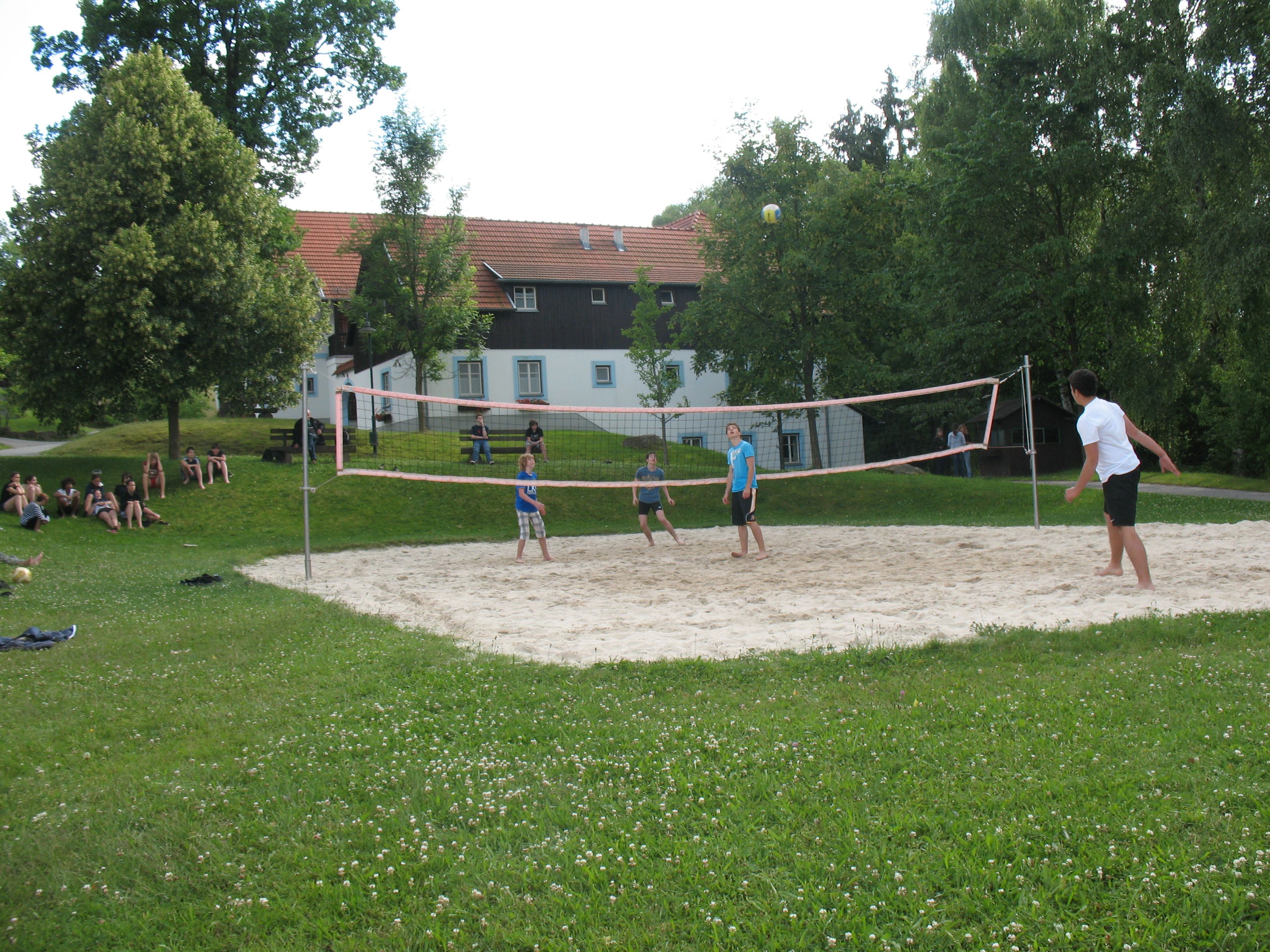 People playing volleyball on an outdoor sand court, surrounded by trees and a building in the background.