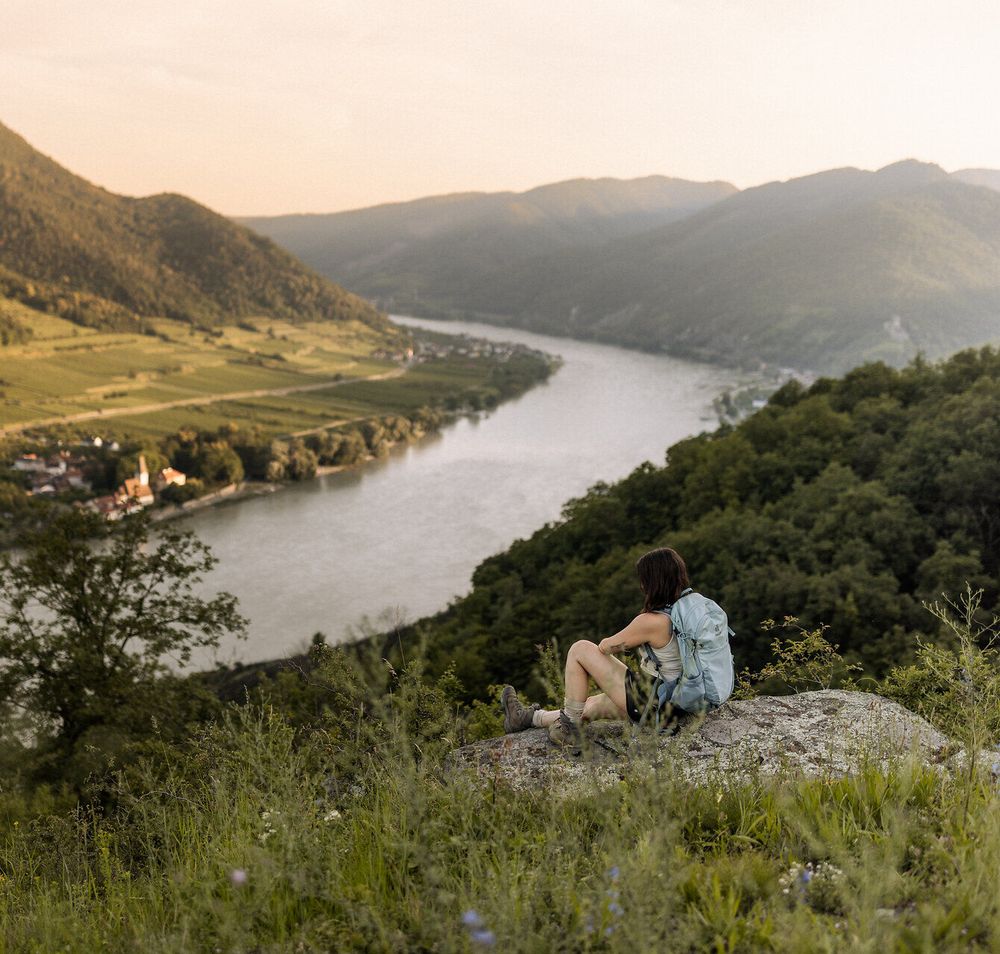 Eine Frau mit Wanderrucksack sitzt auf einem Felsen und blickt auf das Donautal auf Höhe von Spitz an der Donau.