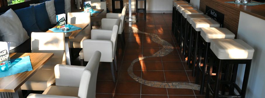 Interior view of a café with white chairs and tables, brown tiled floor and bar stools.