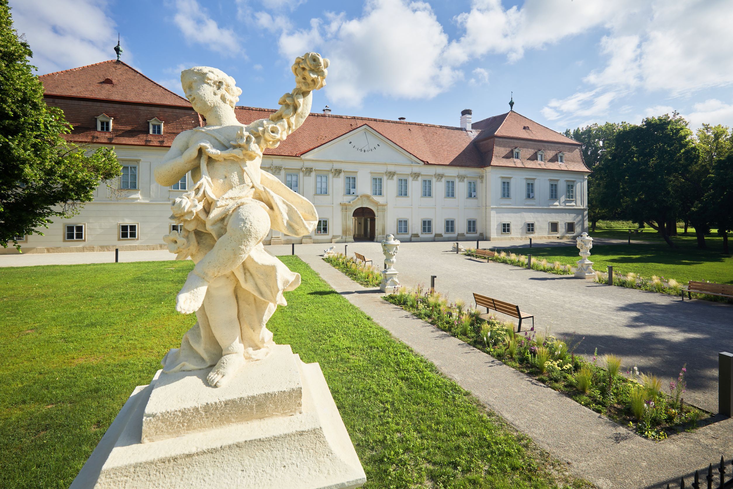Baroque statue in front of a historic building with a red roof and well-tended garden.