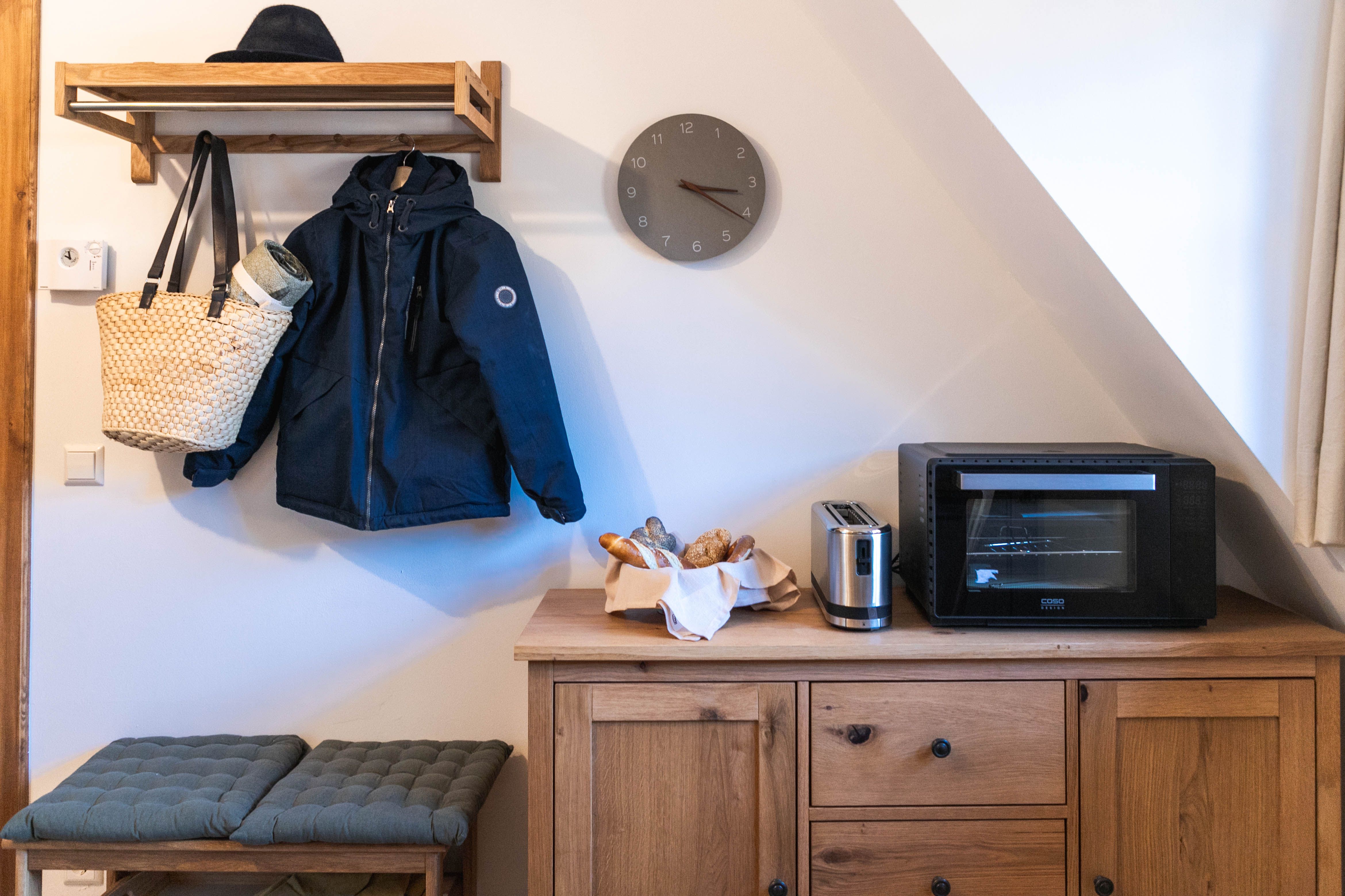 A hallway with a wooden coat rack, a jacket, a hat, a bag, a toaster, an oven and a bread basket on a wooden cupboard.