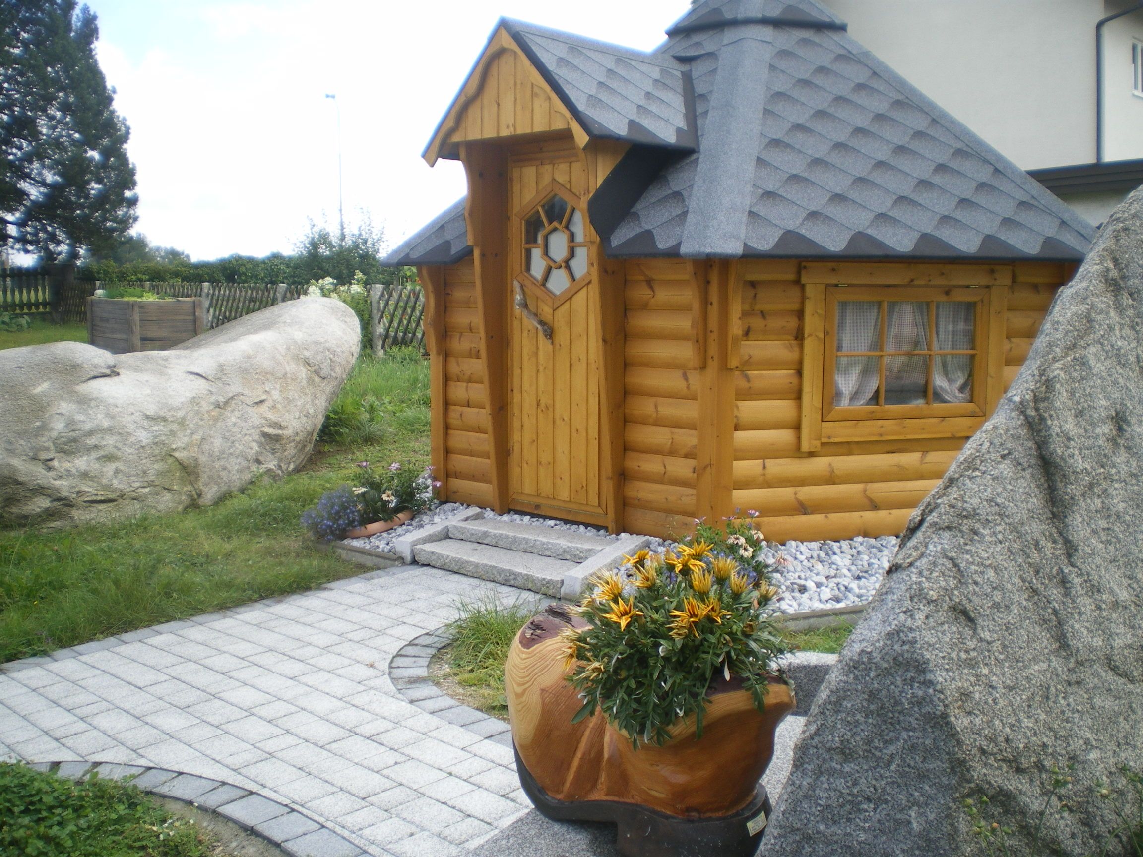 A small, wooden barbecue hut with a gray roof, surrounded by stones and plants.