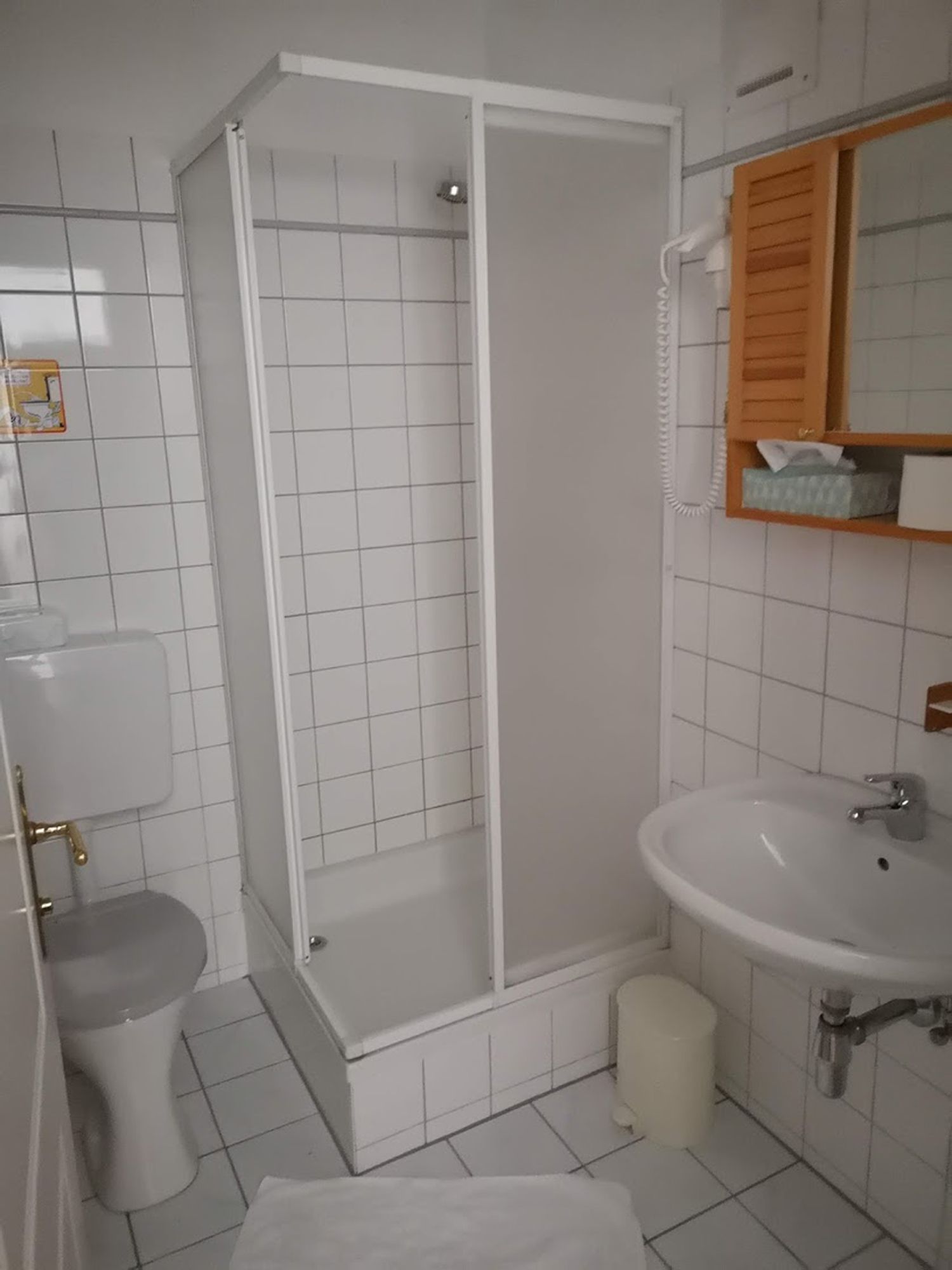 A small, white tiled bathroom with shower, toilet and washbasin.