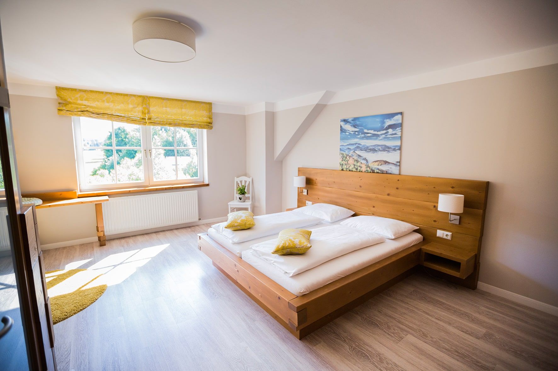 Bright double room with wooden bed, yellow pillows and large window.