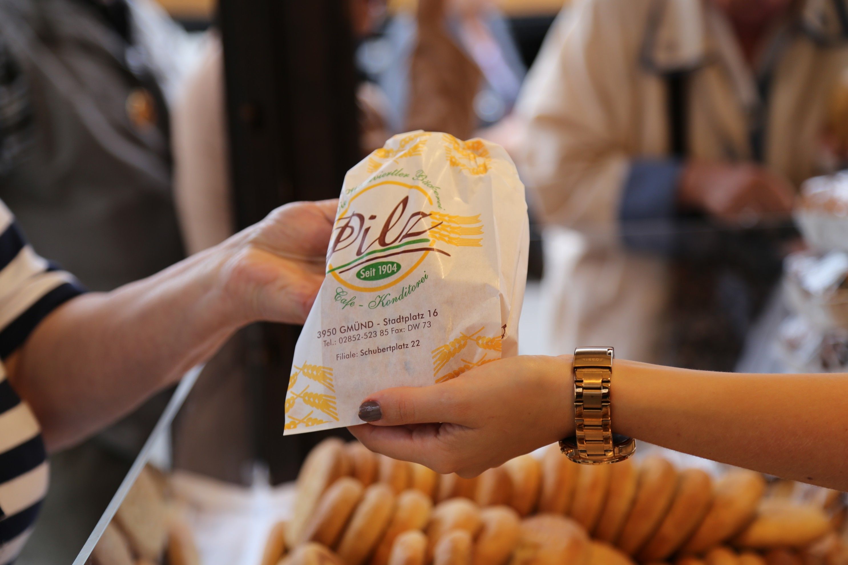 Two hands hand over a paper bag from the Pilz bakery-pastry shop with pastries in the background.