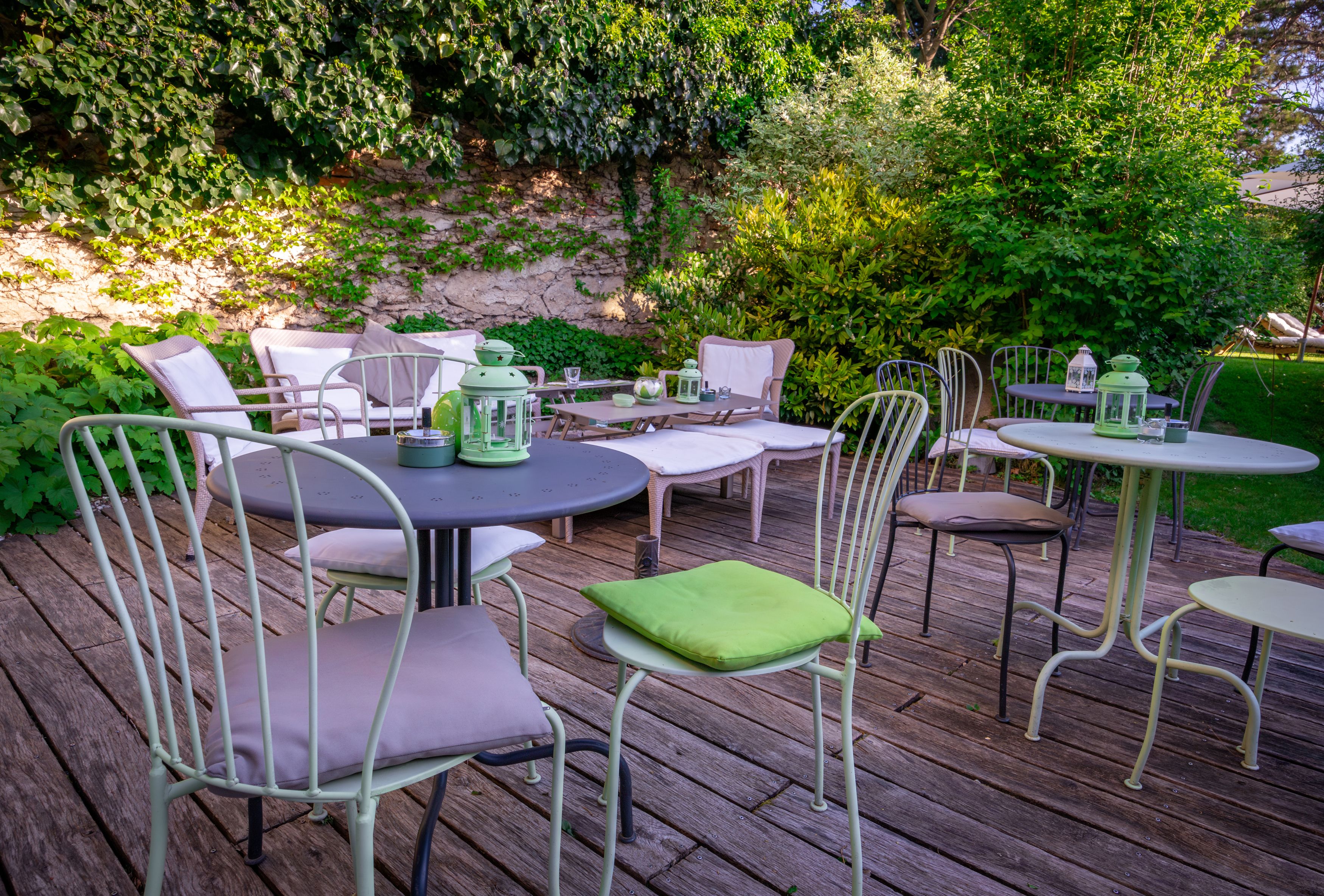 Garden area with tables and chairs on a wooden terrace, surrounded by green plants.