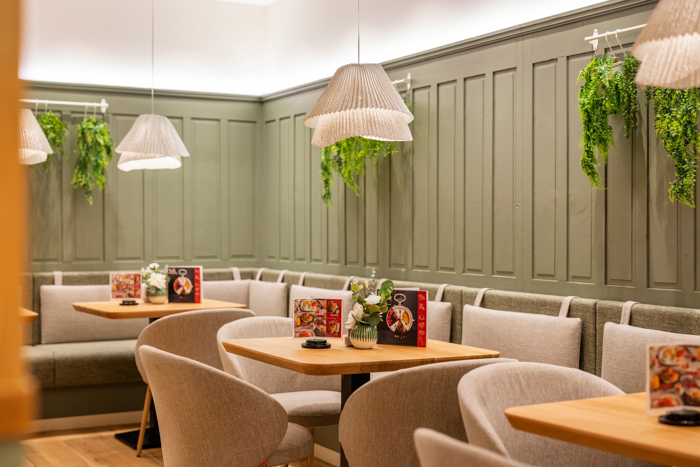 Cozy café area with wooden tables, grey chairs and green wall panelling. Hanging plants and lamps create an inviting atmosphere.