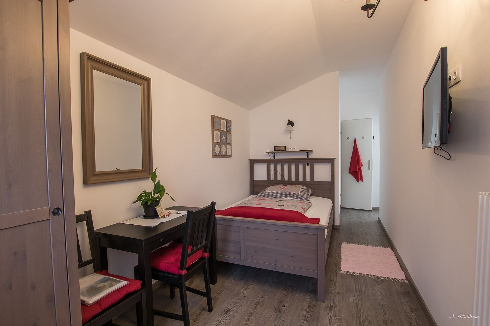 A cozy single room with wooden furniture, a bed, a small table with chairs, a mirror and a TV on the wall.