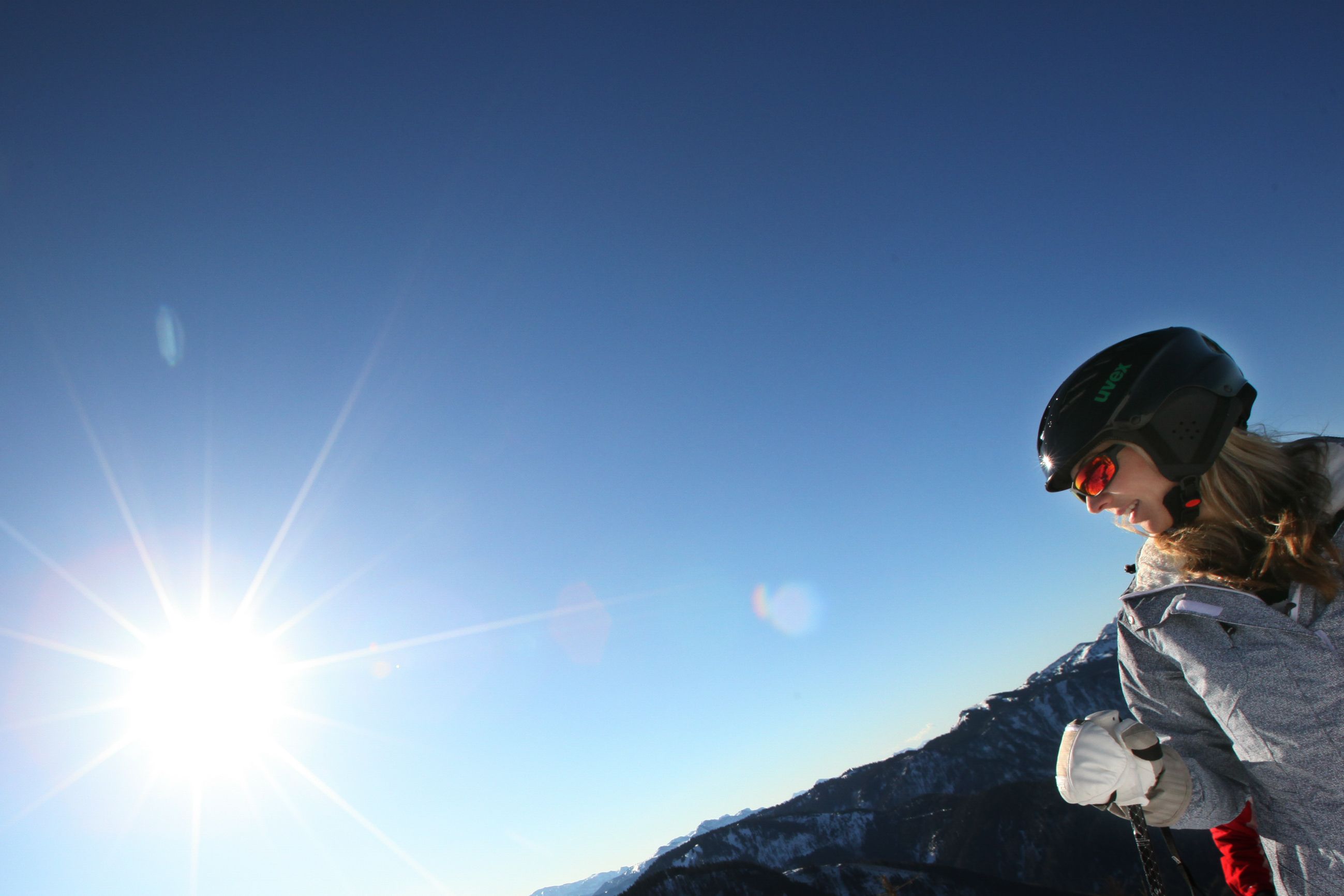 A person wearing a helmet and ski goggles stands in front of a sunny, clear sky in a mountain landscape.